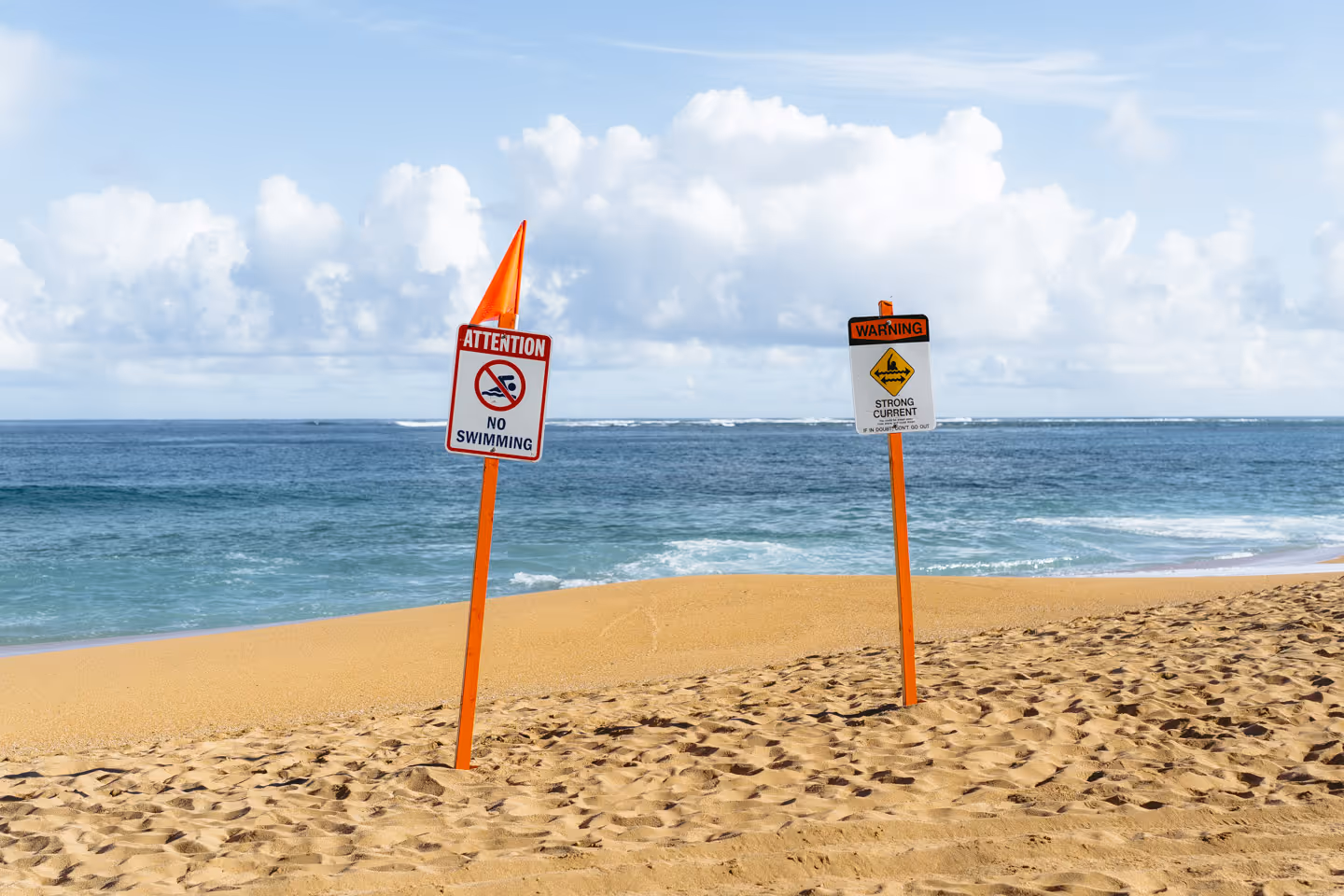Two warning signs on a sandy beach near the ocean, one cautioning swimmers, with waves and clouds in the distance.