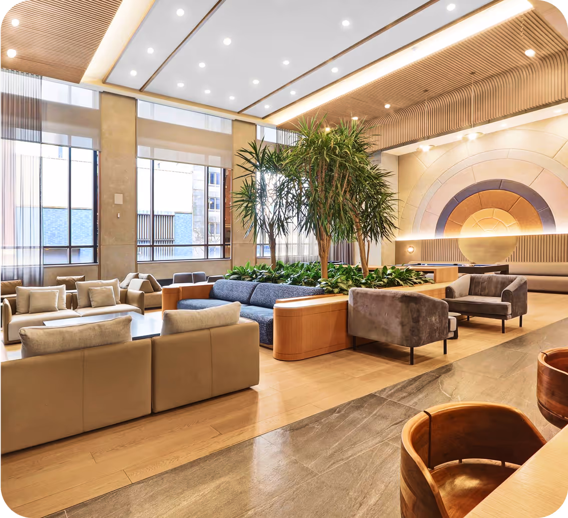 Modern lounge area with beige and gray sofas, indoor plants, large windows, and decorative circular wall art.