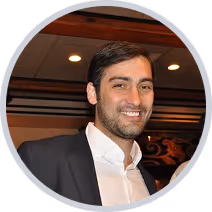 Smiling man with dark hair wearing a white shirt and dark blazer in an indoor setting.