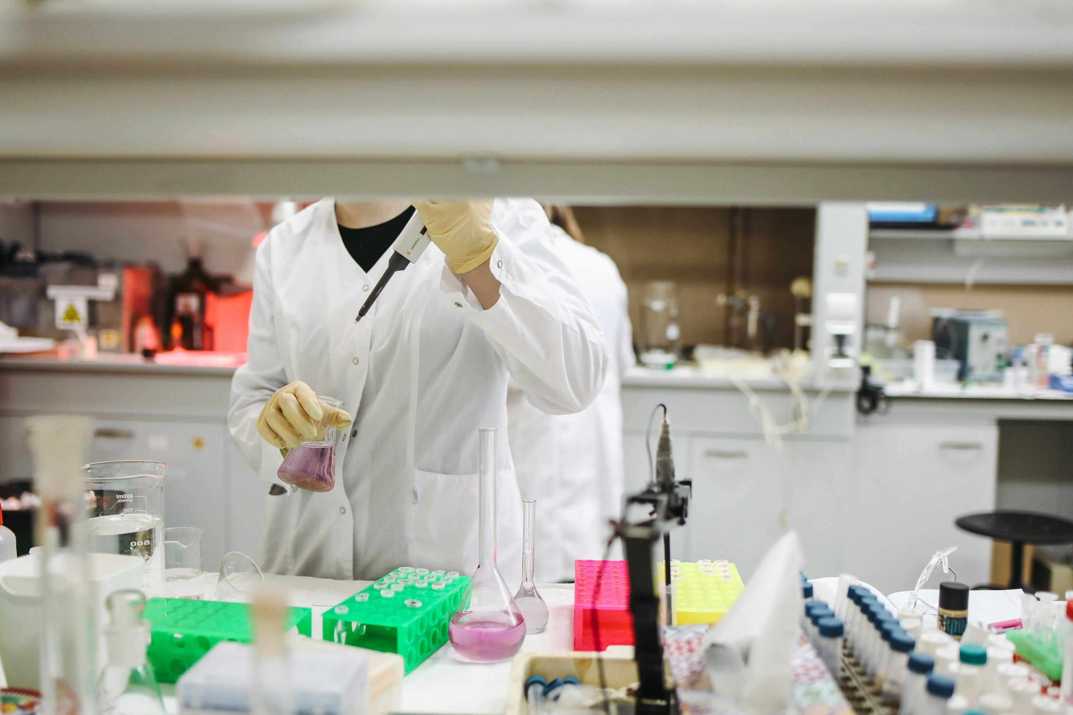 A lab worker in a white coat uses a dropper to add liquid to a beaker with purple fluid.