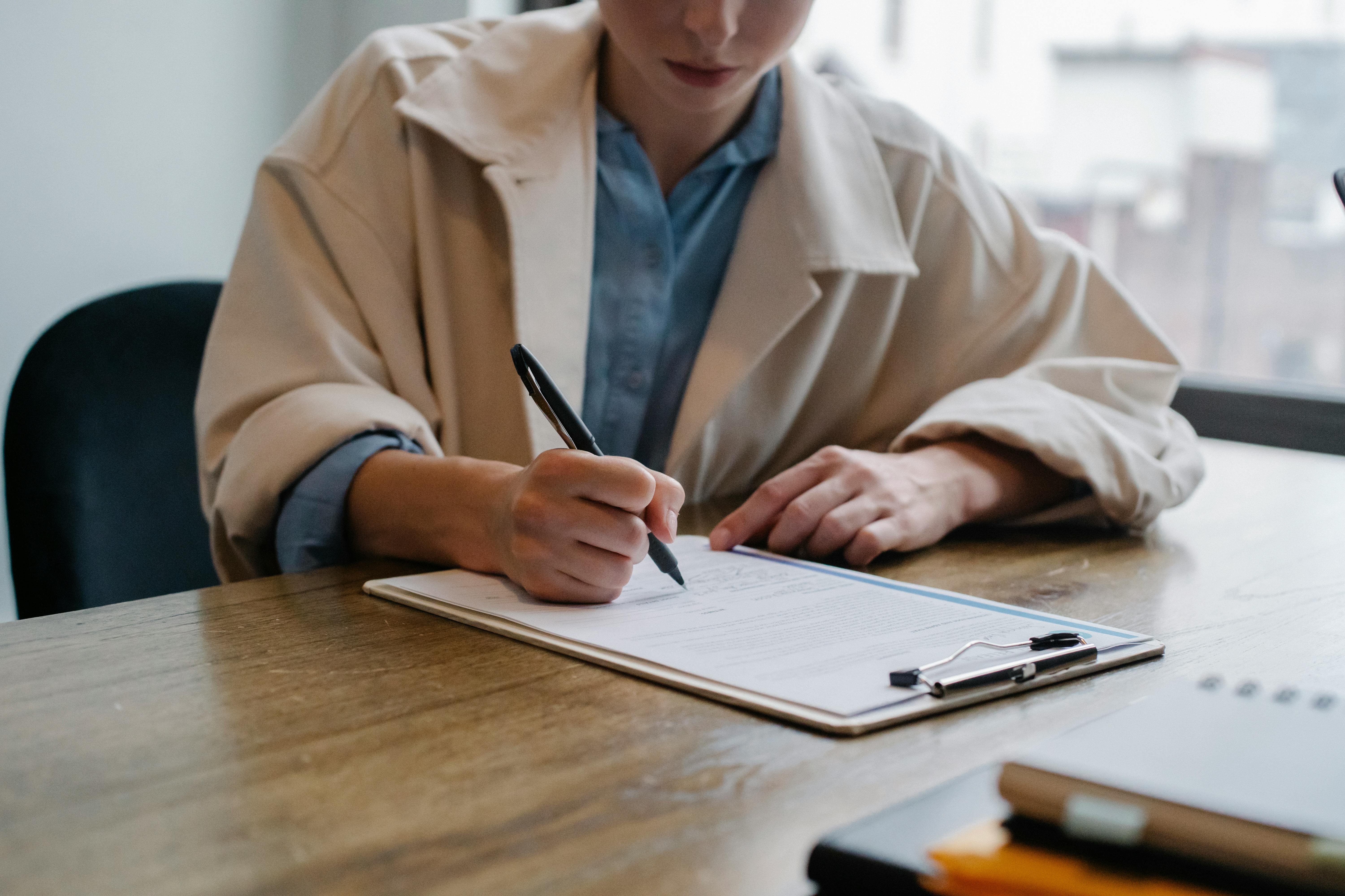A person wearing a light coat sits at a wooden desk, writing on a clipboard with a pen. 