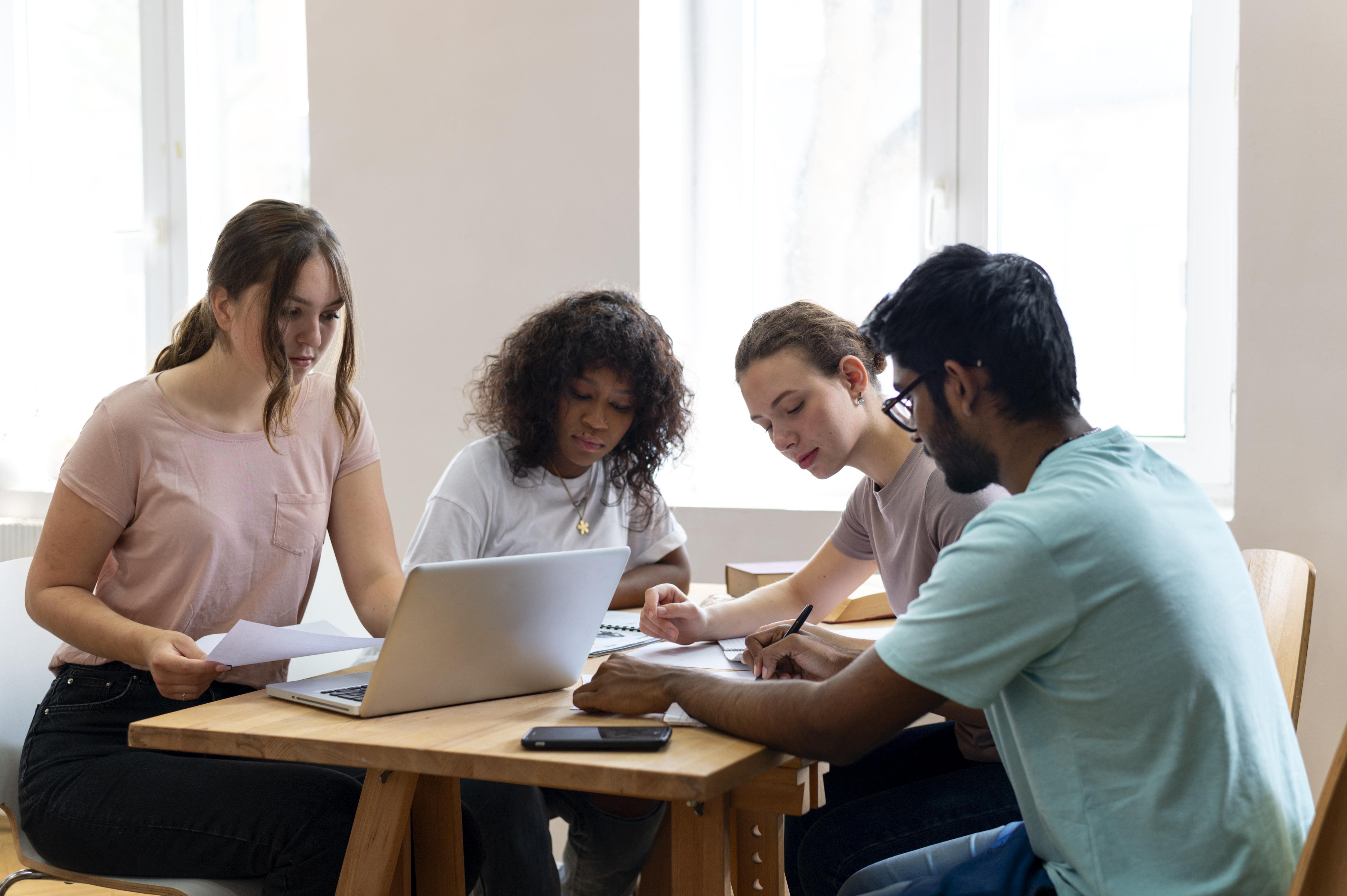 Four people from different backgrounds sit at a wooden table. They work together using a laptop, papers, and pens in a well lit room.