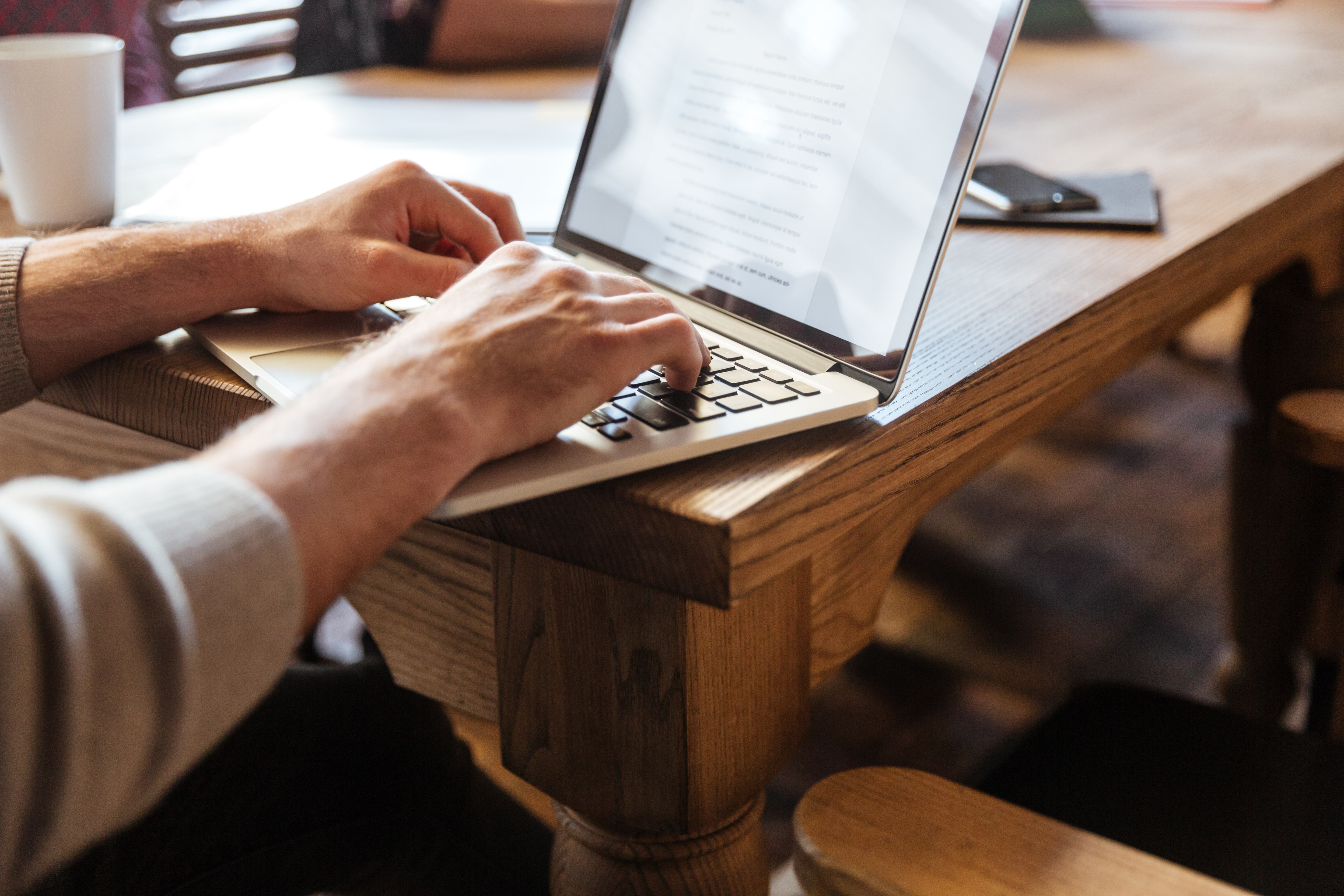 A person types on a laptop at a wooden table. A coffee cup and phone sit nearby.