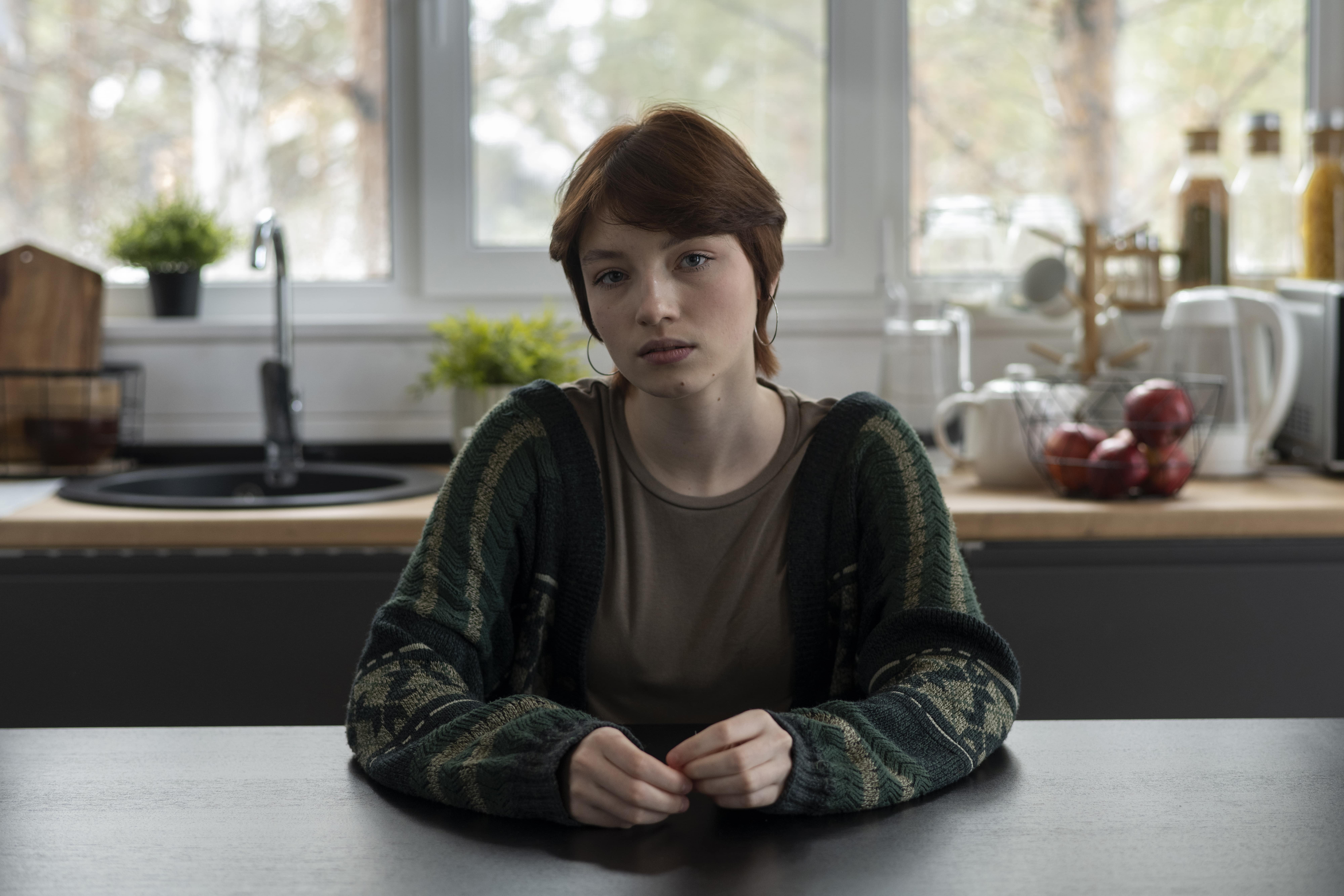 A young woman sits at a table staring at the camera with a serious face.