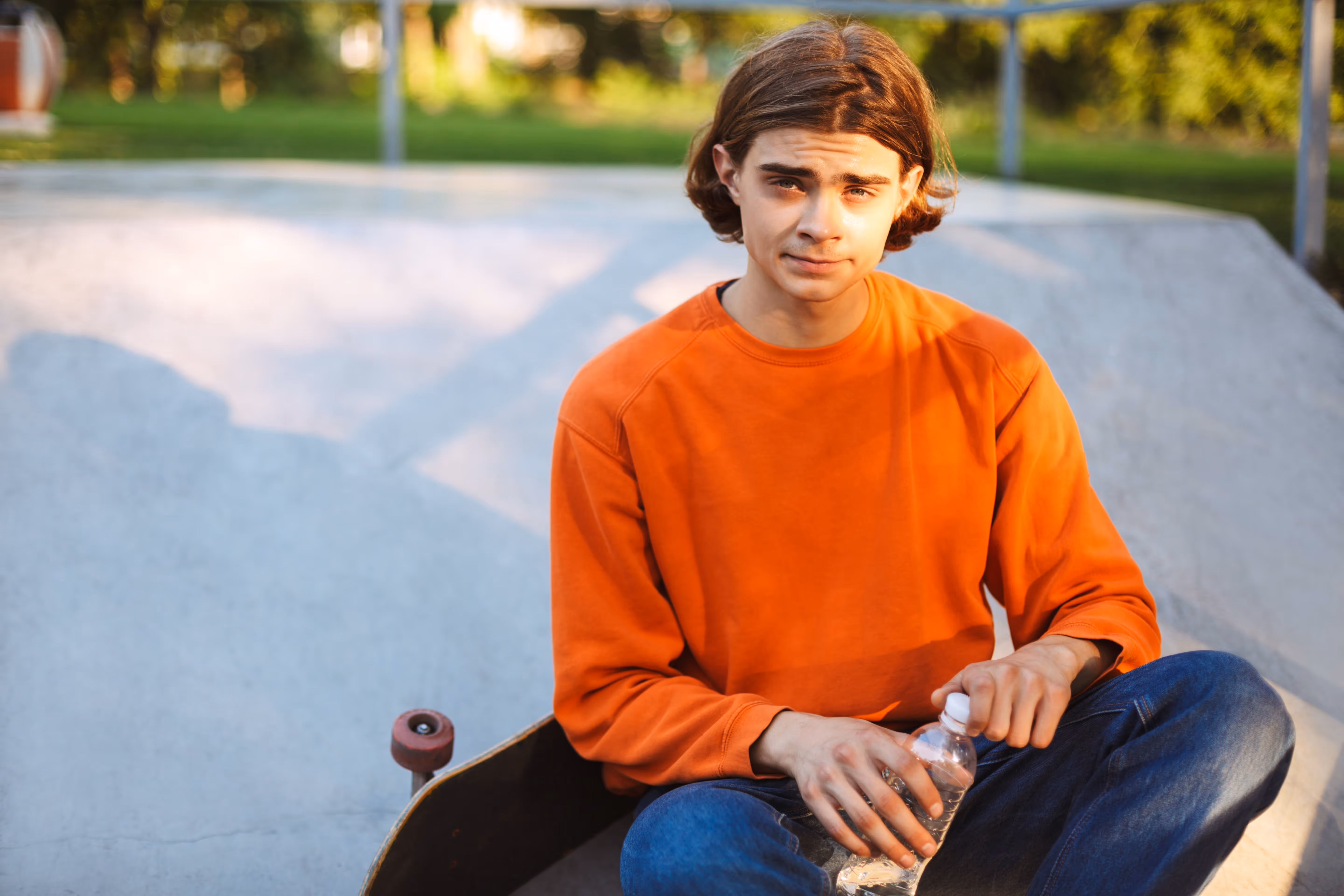 A person in an orange sweatshirt sits on a skateboard at a sunny skatepark.