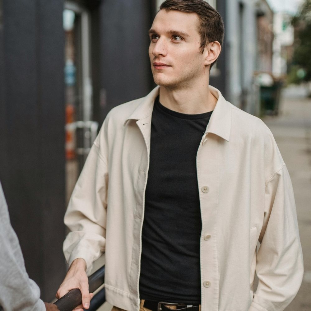 Guy in beige jacket stands on a sidewalk engaged in conversation.