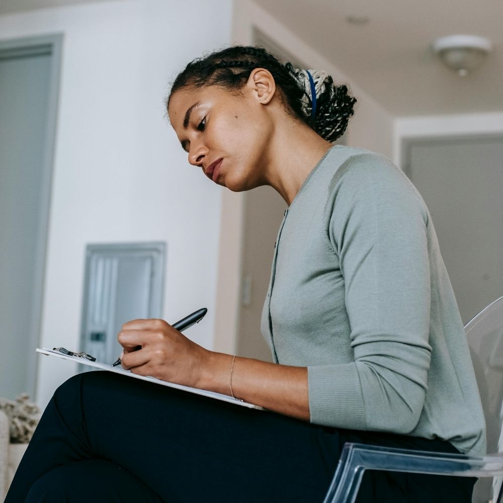 Woman writes in notebook on transparent chair in modern, stylish living space.