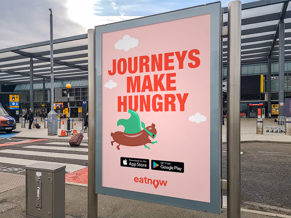 A pink billboard outside an airport with bold red text that says "Journeys Make Hungry" above a sausage brand mascot wearing a green superhero cape, promoting the eatnow food app.