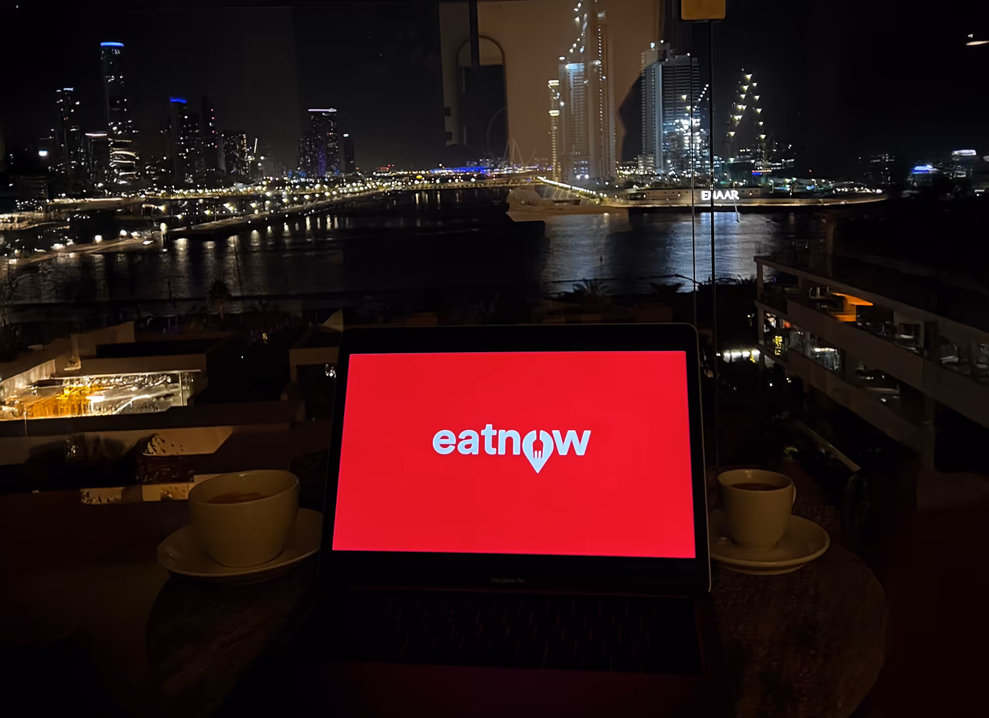 Laptop displaying the red eatnow logo on screen, placed on a table with two coffee cups, overlooking the Dubai city skyline and waterfront at night.