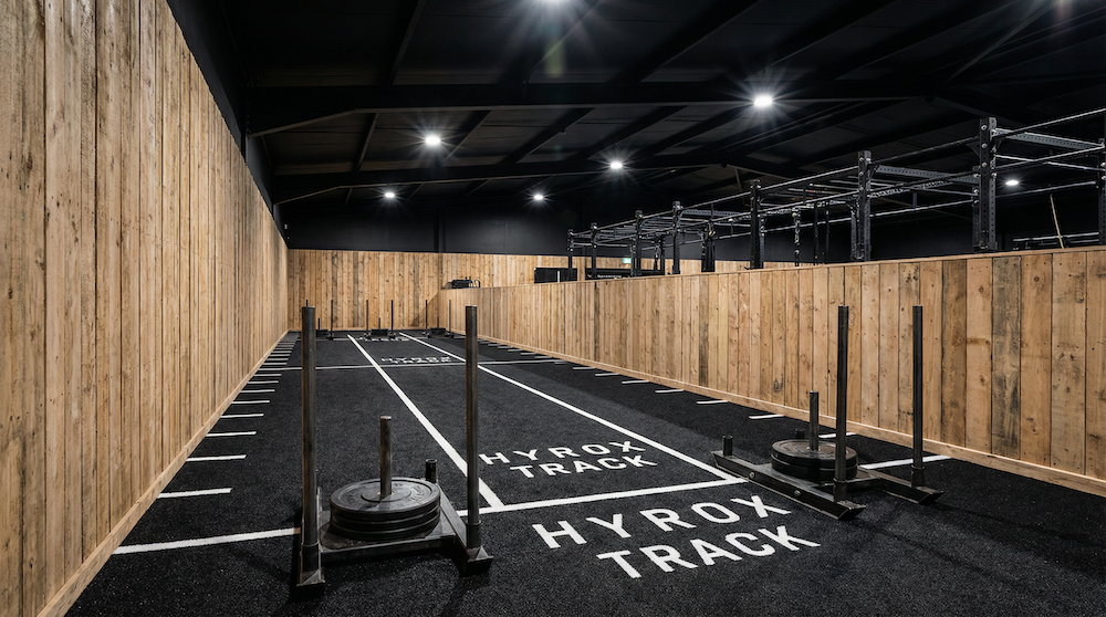 Indoor gym sled track with weights on two sleds on black flooring marked with 'HYROX TRACK' and wooden paneled walls.