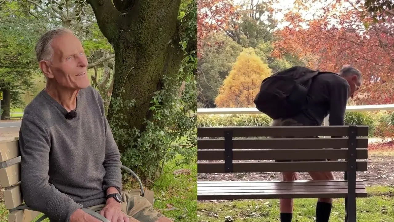 Elderly man sitting then standing from a park bench surrounded by trees in a leafy outdoor setting.