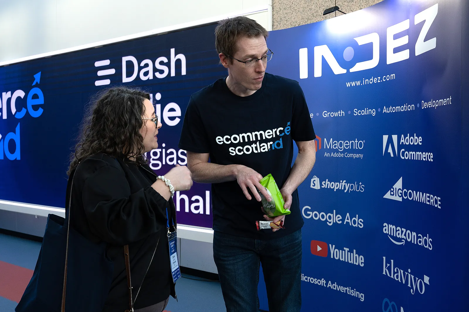 Two attendees chatting at the INDEZ stand in front of the Ecommerce Scotland 2026 sponsor wall