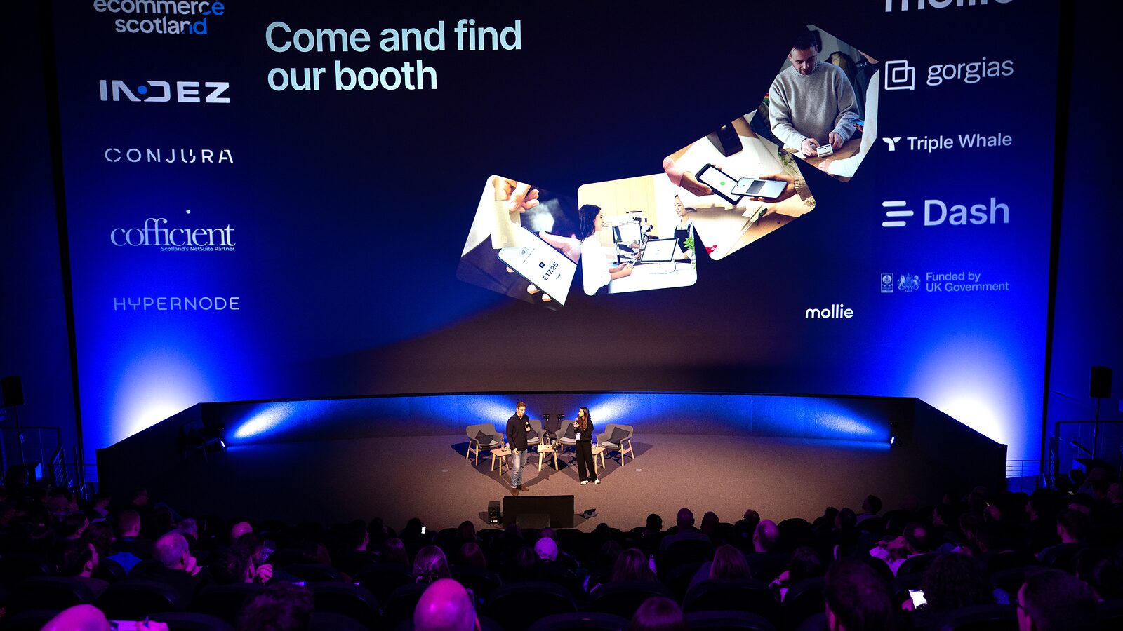 Mollie’s “Come and find our booth” slide on the IMAX screen, with two people on stage and the audience in the foreground