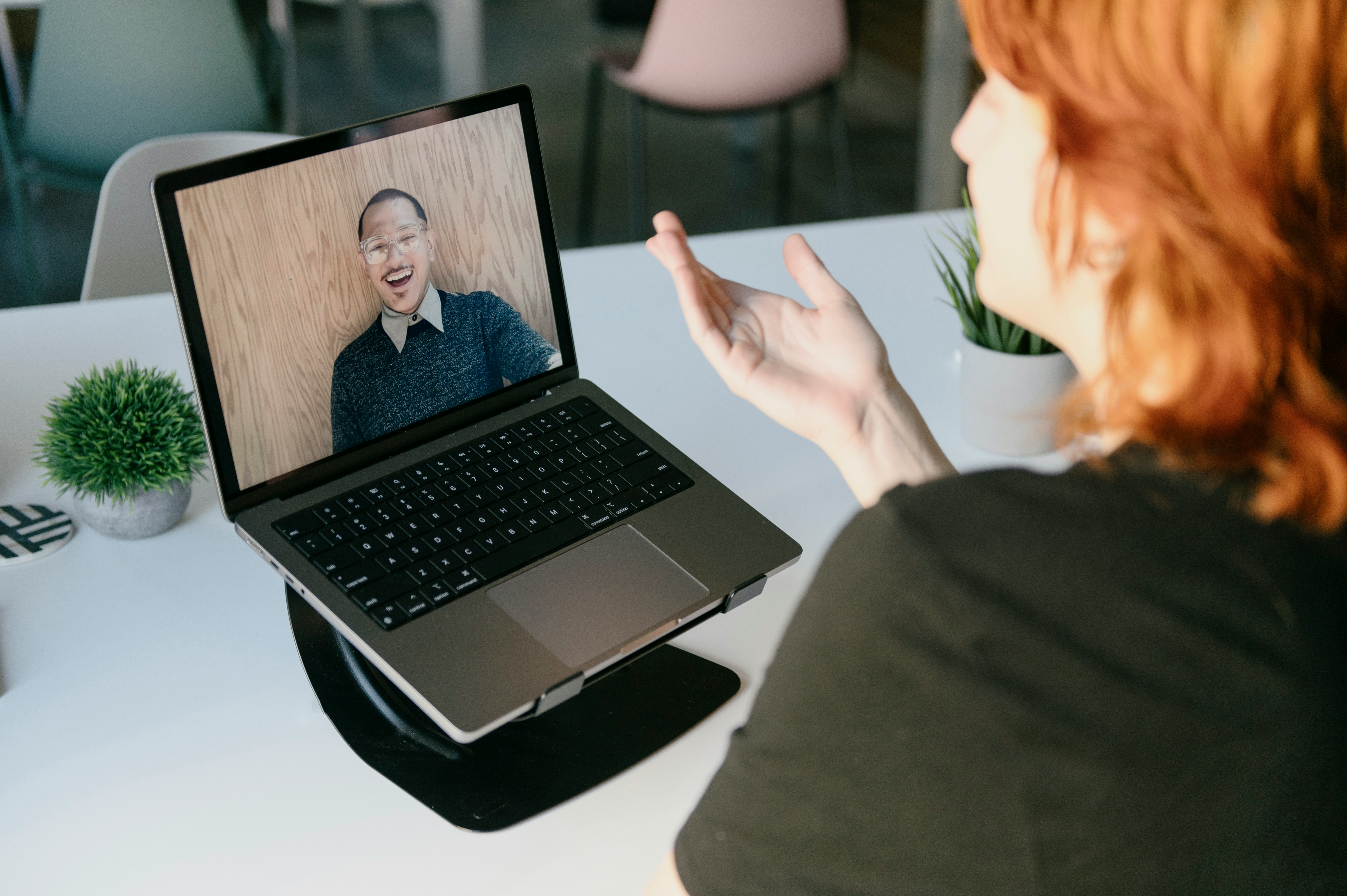 Person with red hair engaging in a video call on a laptop showing a smiling man with glasses in front of a wooden background.