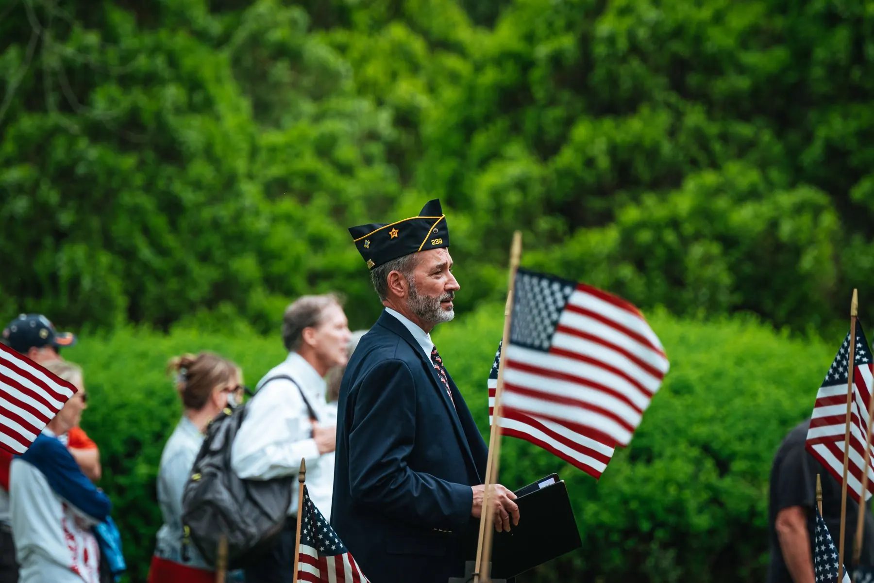A veteran in a service cap holds an American flag at an outdoor Veterans Day parade