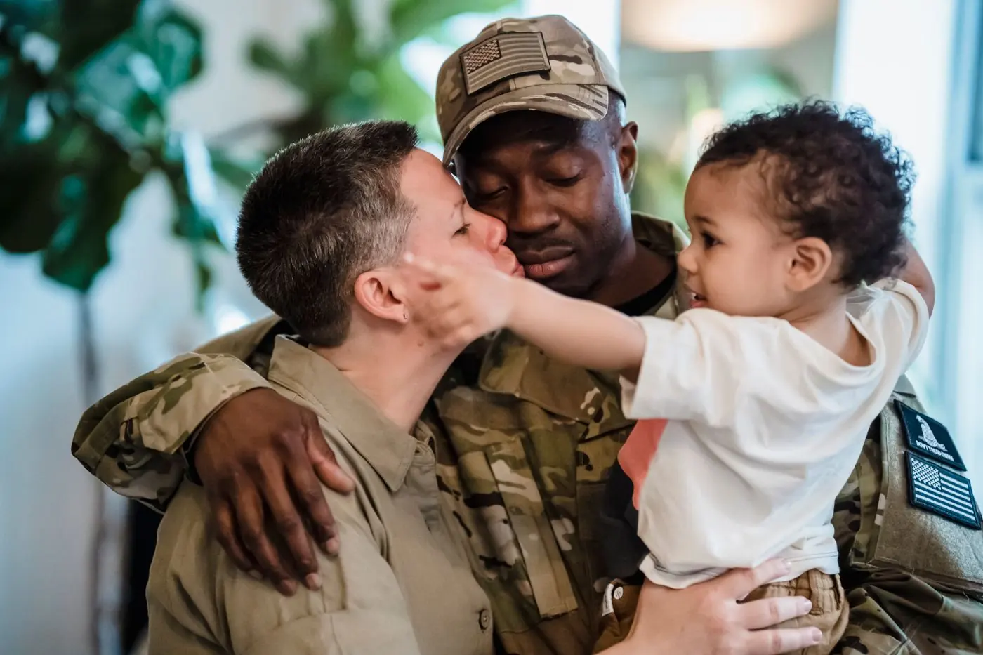 A service member in uniform embraces their partner and young child at home