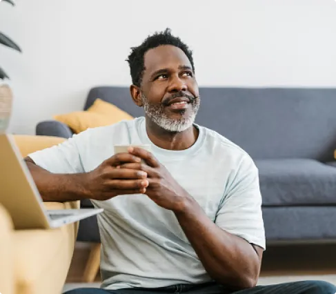 Man sitting on the floor holding a white cup, looking thoughtfully to the side with a laptop open beside him.