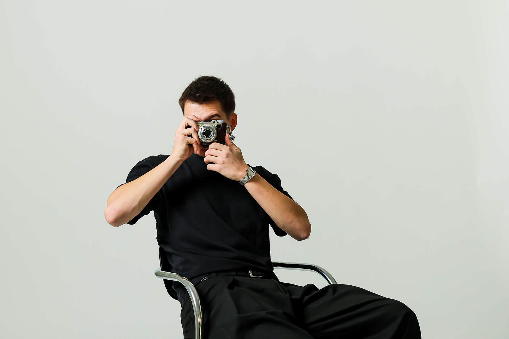 Man with beard wearing a brown sweater sitting thoughtfully on a chair against a plain light background.