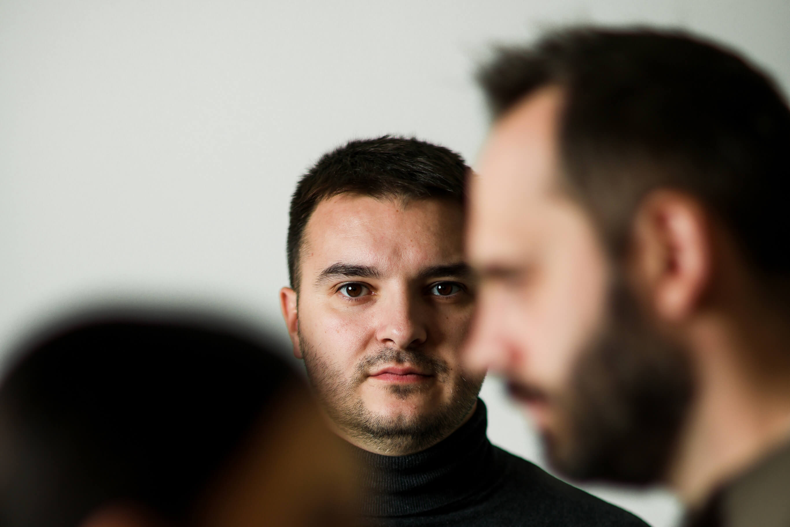 Bearded man in a brown sweater sitting thoughtfully on a black chair against a plain light background.