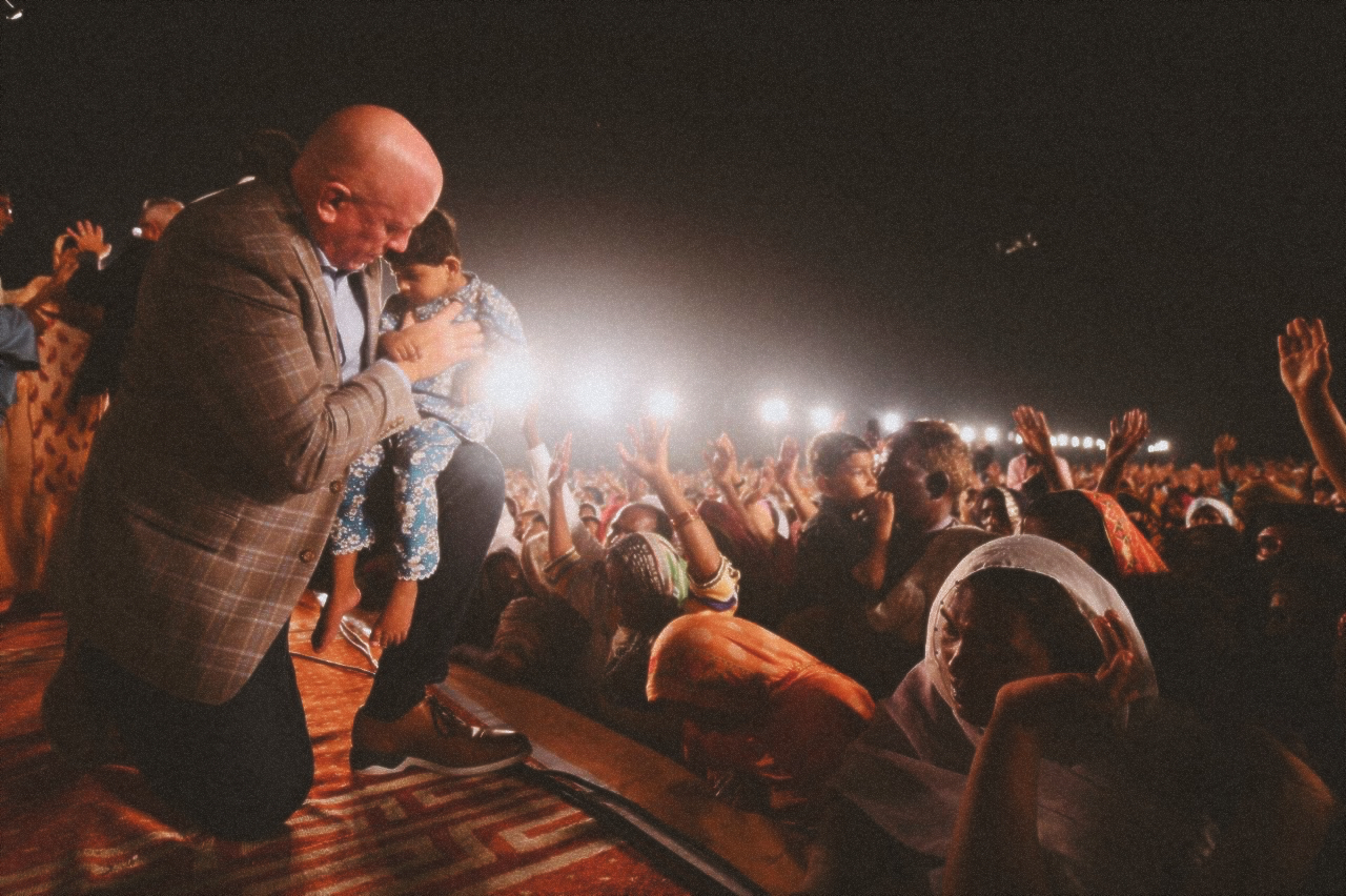Pastor David on stage at a crusade in Pakistan, holding a young crippled boy in his arms, praying. 