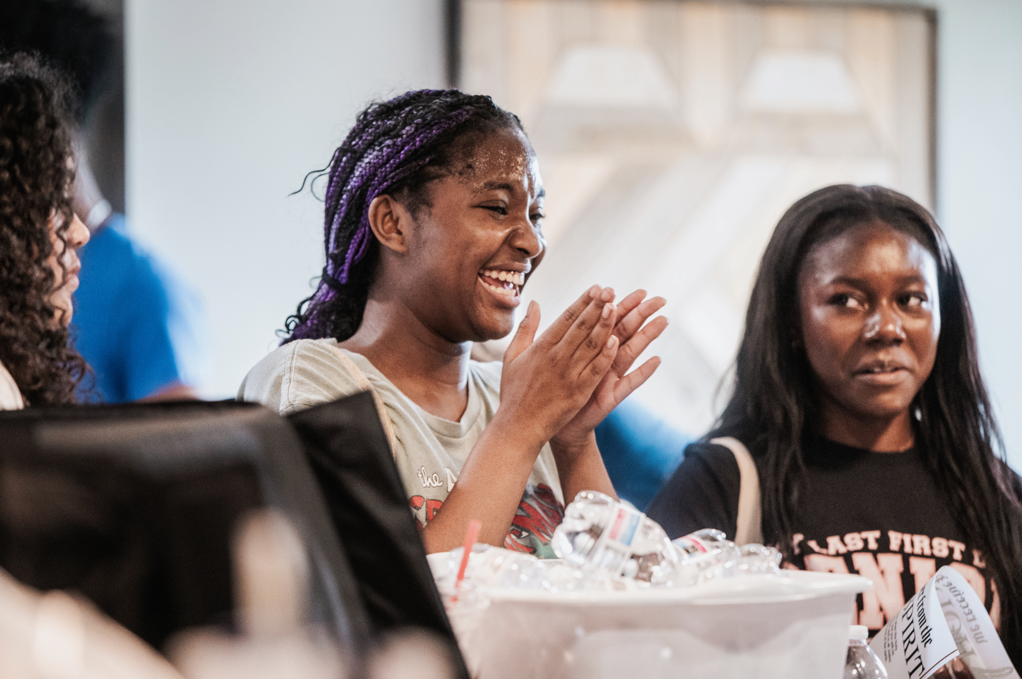 A group of of young women interacting and smiling, one girl claps her hands happily. 