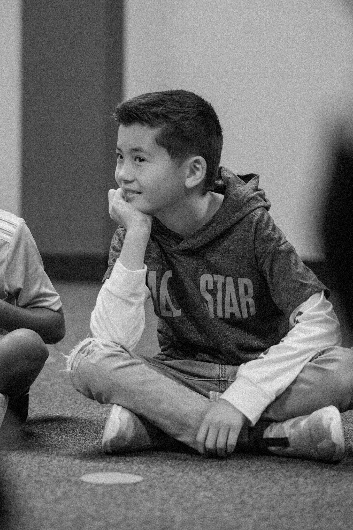 A black and white image of a young man sitting cross legged on the floor in a circle of other Harvest Kids during a lesson