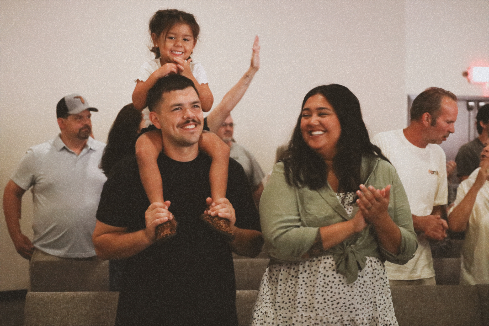 An image of a young couple smiling and enjoying worship at the Independence campus. The young man has his little girl on his shoulders, his wife smiles as she claps and looks at them adoringly.