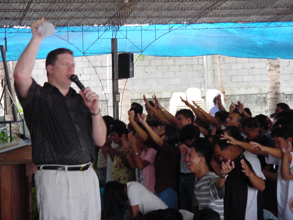 Pastor David holds a microphone while a large crowd raises their hands in worship during a covered church service.