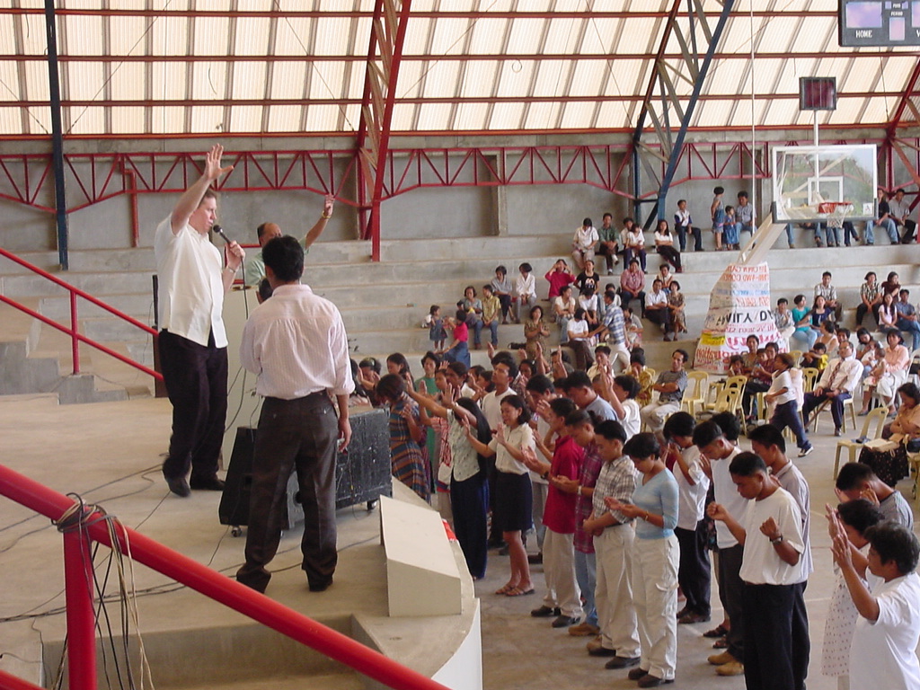 Pastor David gestures while preaching in a building with red steel beams, addressing attendees seated on concrete steps.