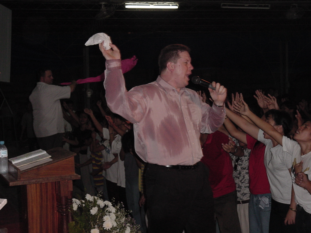 Pastor David preaching passionately while the congregation raises their hands in prayer and worship under a covered structure.