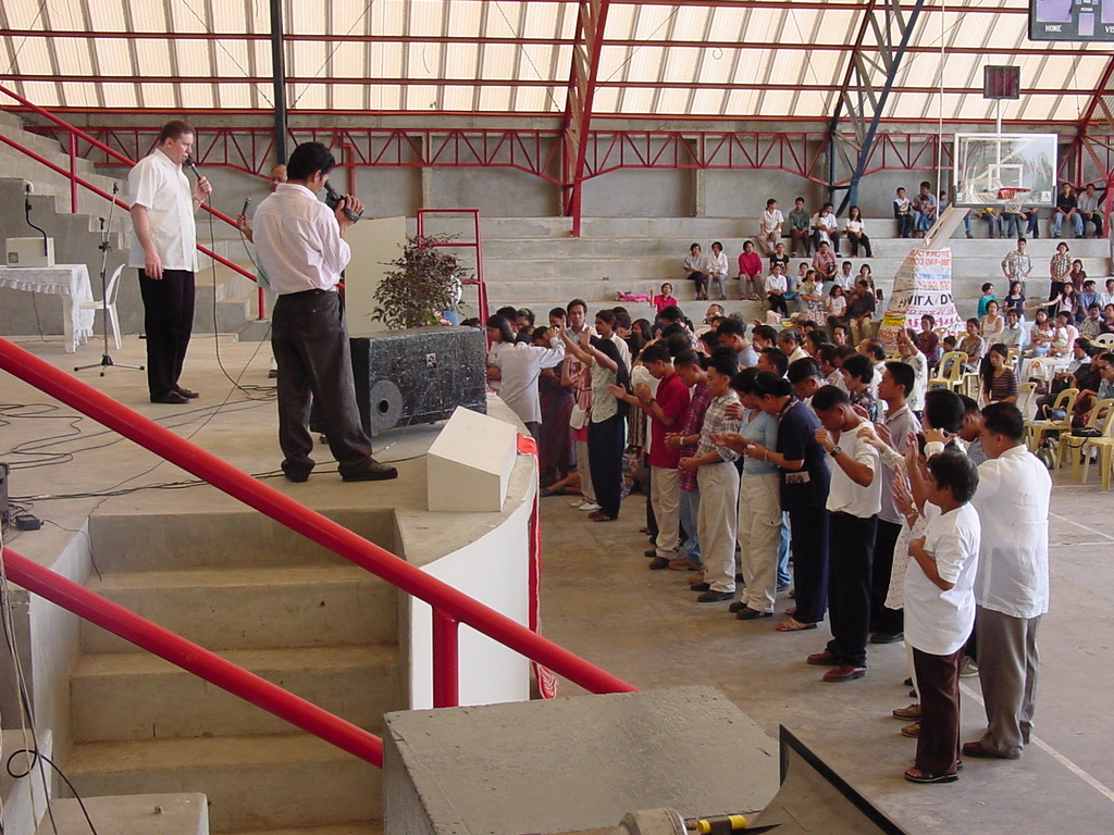 Pastor David leads an alter call to a large group gathered near the altar while another man interprets beside him in a crowded indoor meeting.
