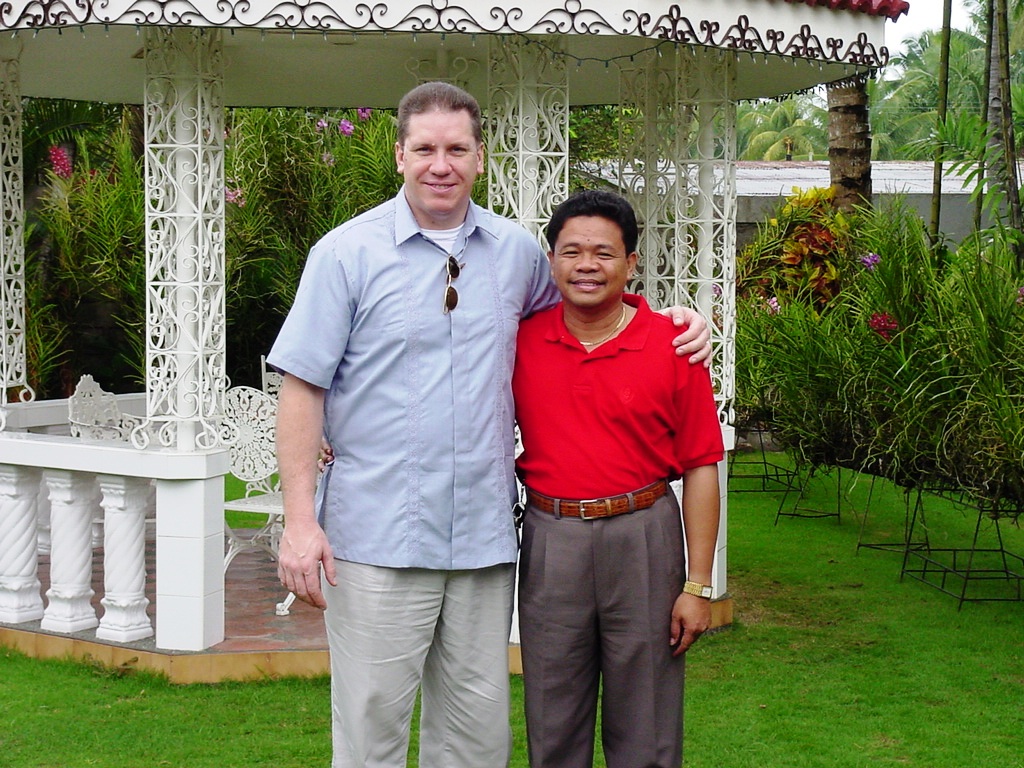 Pastor David and Tony stand arm-in-arm smiling for a photo in front of a white gazebo surrounded by lush tropical plants.