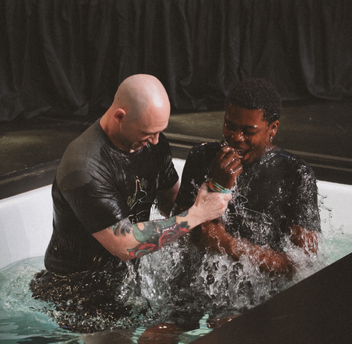 Two men in black shirts performing a baptism with water splashing inside a pool.