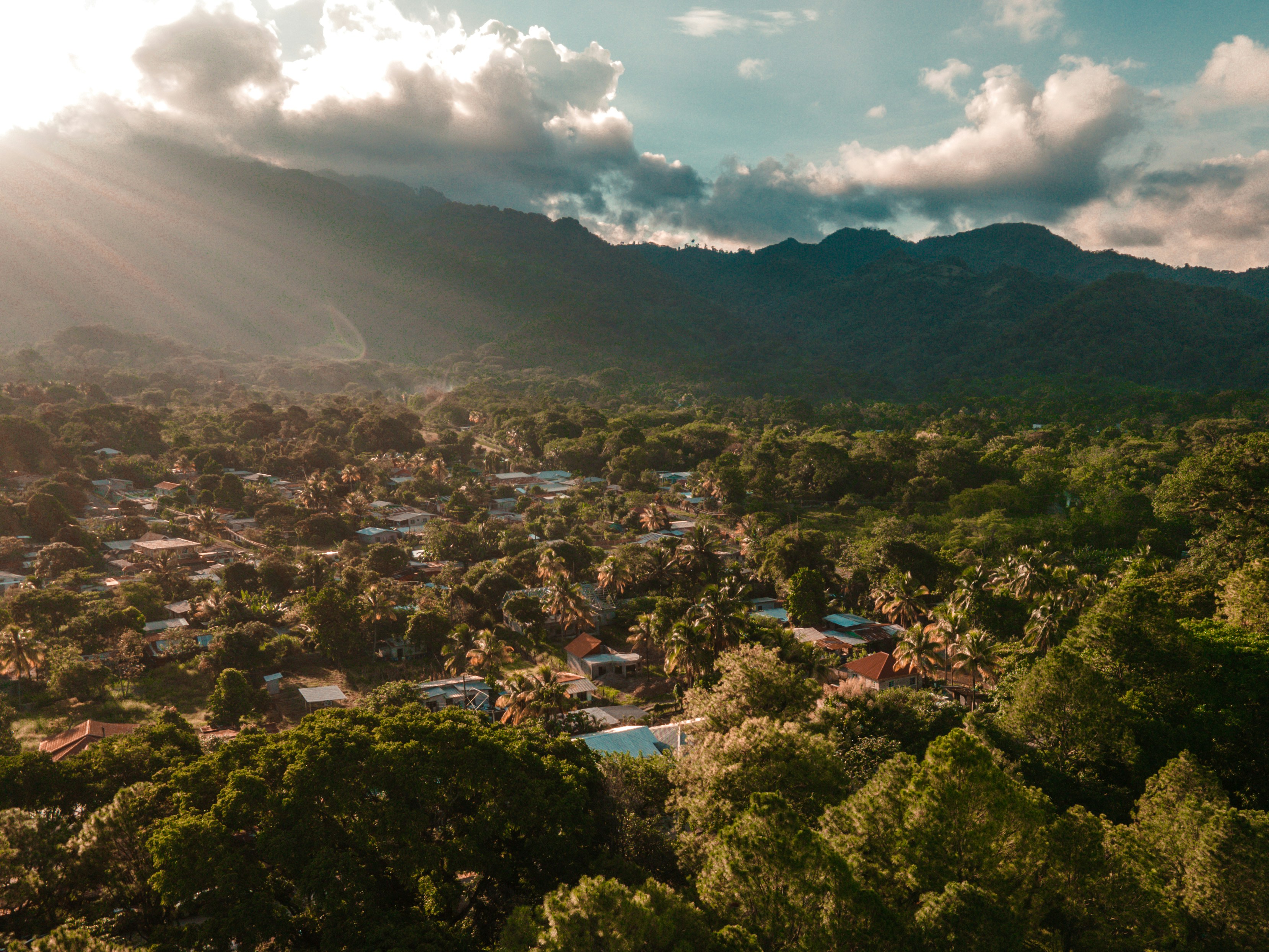 Sunlight breaking through clouds over a lush tropical village surrounded by dense trees and mountains in the background.