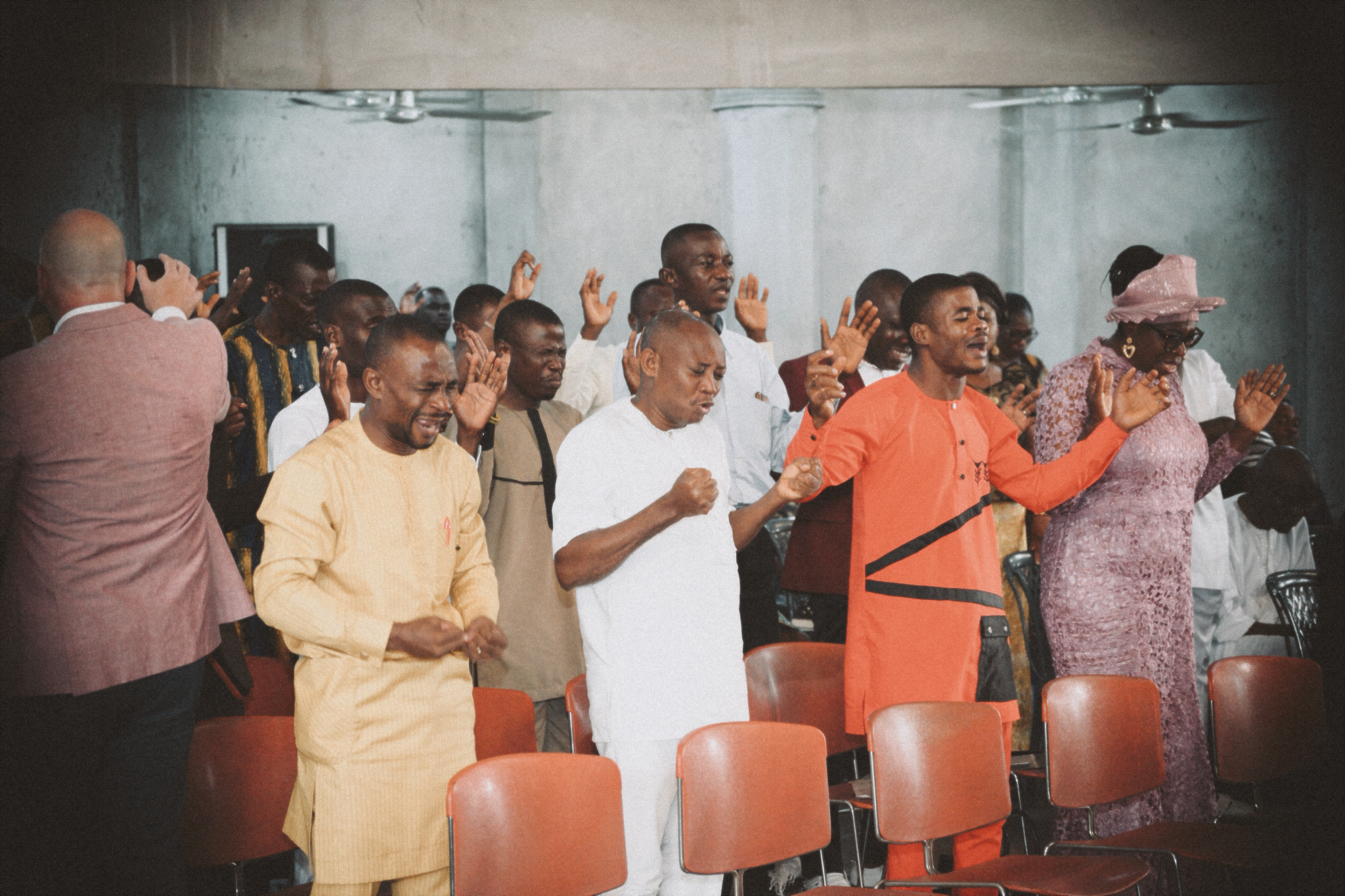 A group of men worshiping during a church service as Pastor David prays over members of the congregation in the background.