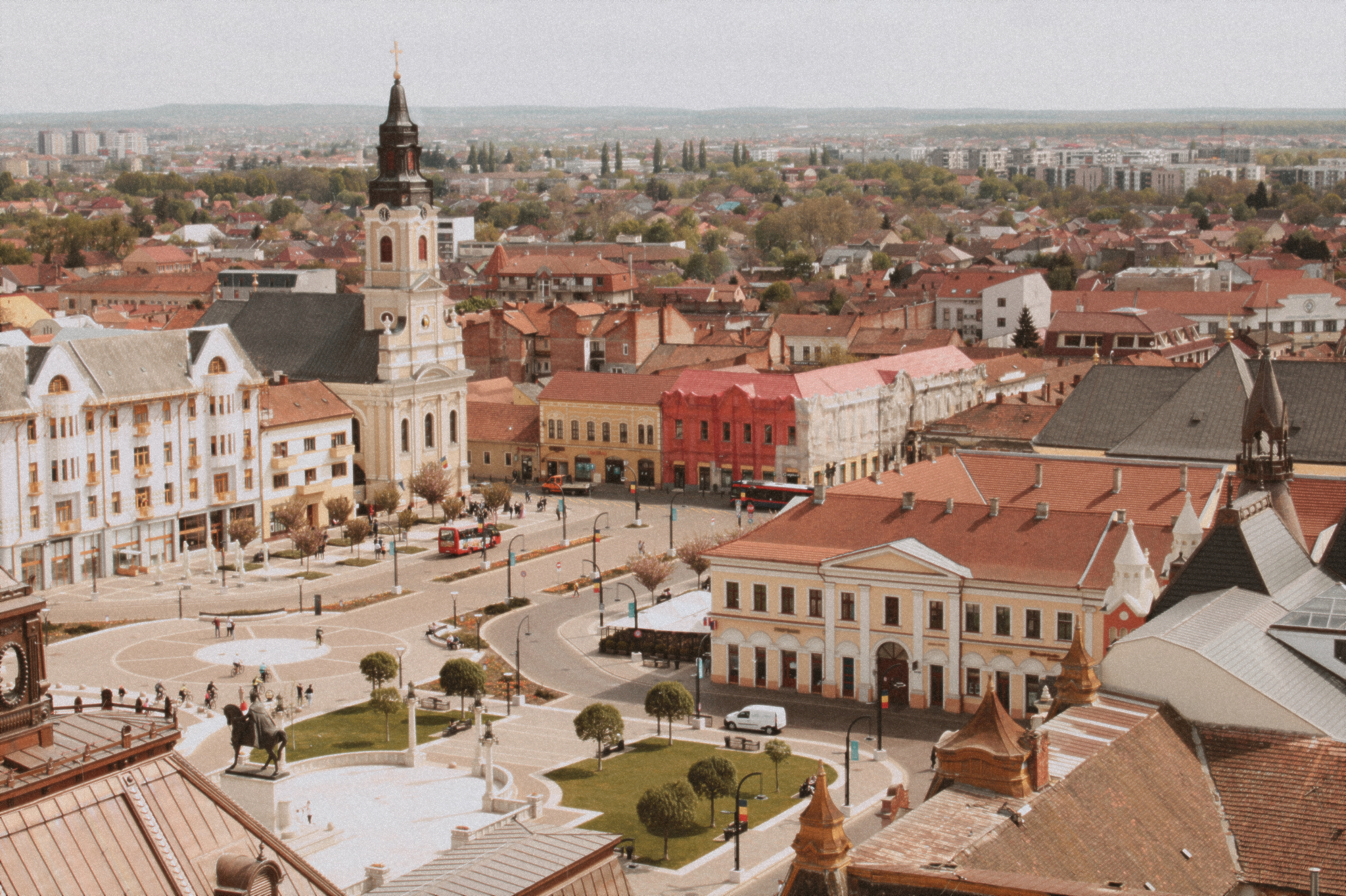An Aerial view of a beautiful Romanian city square. The buildings have red roofs, and there is a cream chapel and its black steeple standing prominently in the foreground. 