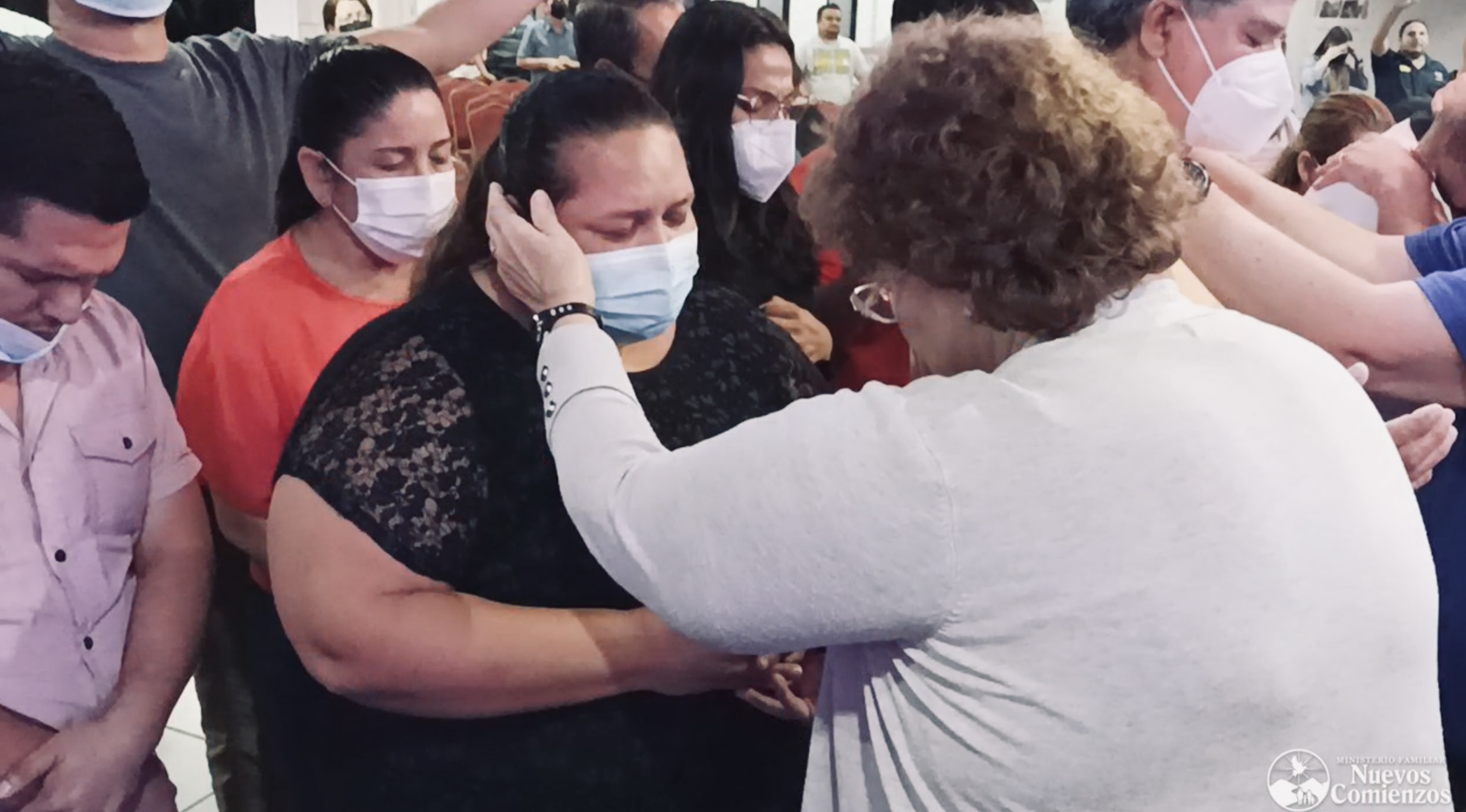 A woman in a white sweater lays her hand on the face of a woman in a mask in prayer