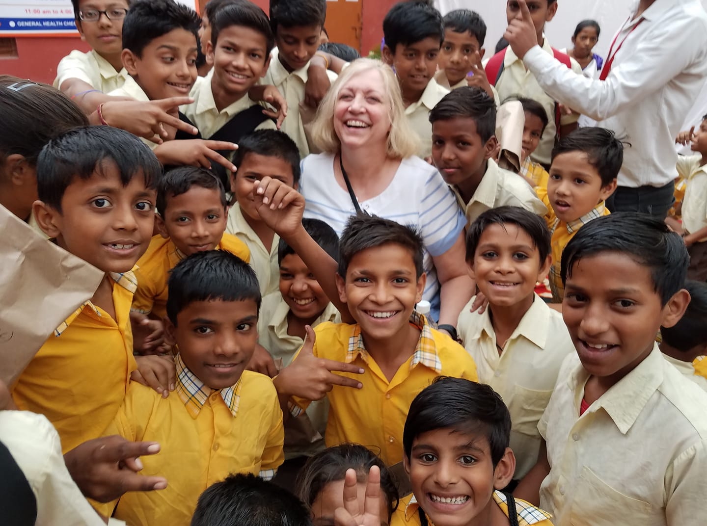 A cheerful group of schoolchildren in yellow shirts crowd around a smiling woman, laughing and posing together for a joyful photo at a school event.