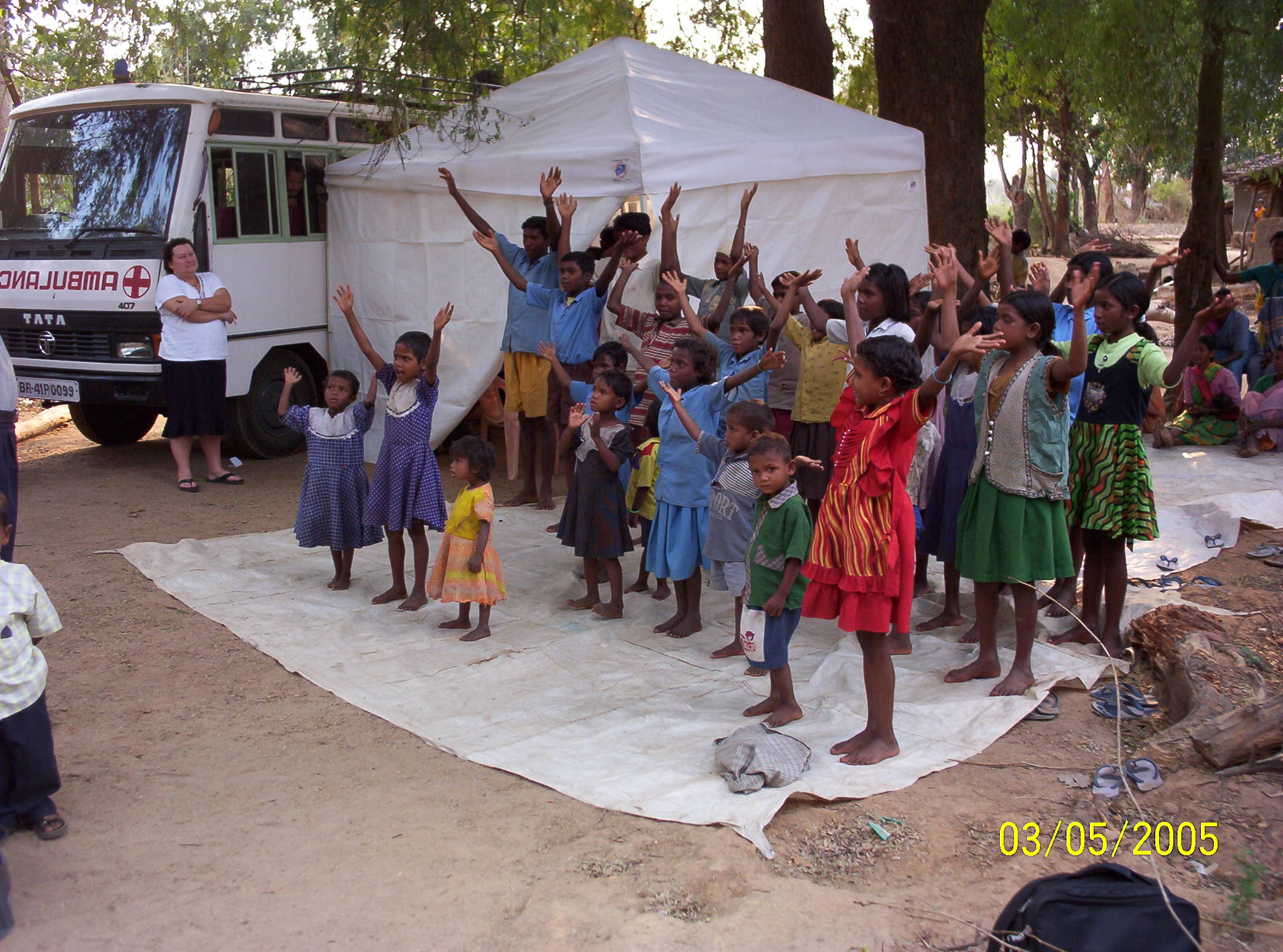 A group of children stand with their hands raised during an outdoor gathering near an ambulance and tent, as adults observe in the background.