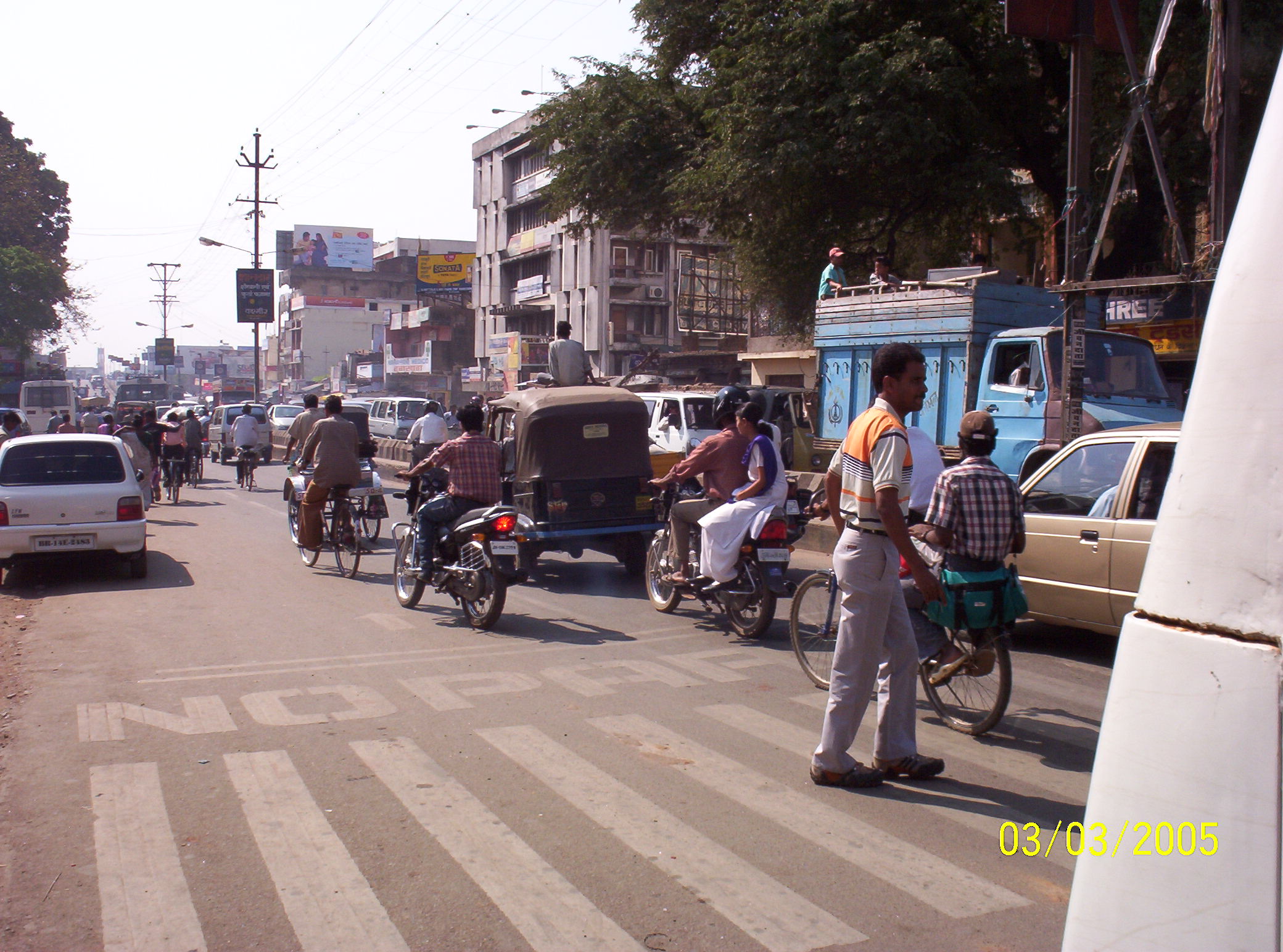 A busy urban street in India with cars, motorbikes, and cyclists moving through morning traffic as pedestrians walk along the roadside.
