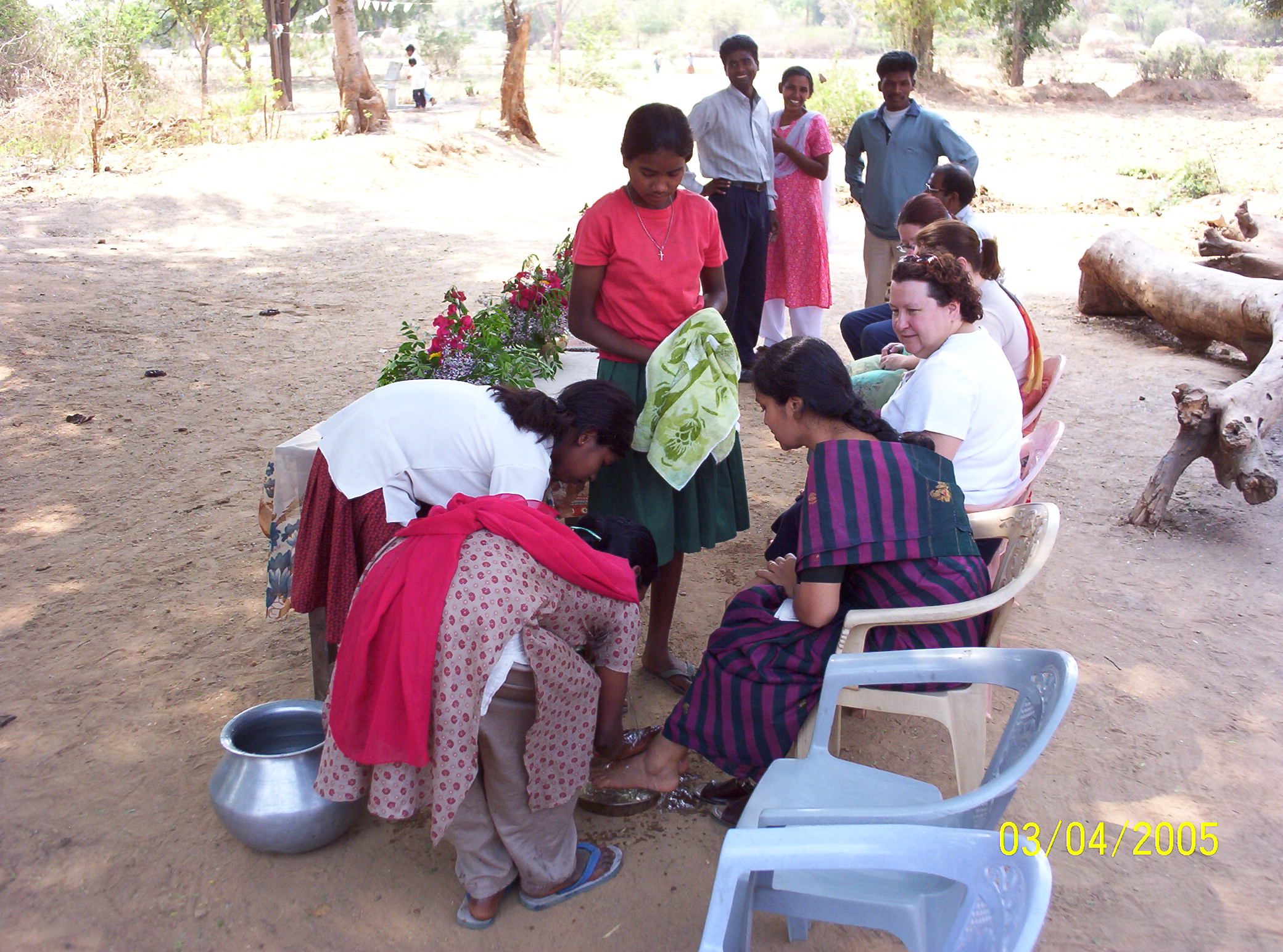 Young girls wash the feet of seated women outdoors, surrounded by onlookers and trees, as part of a cultural or religious ceremony.