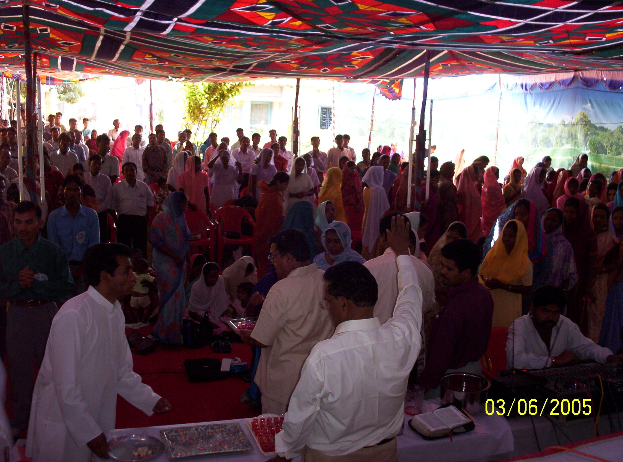 A large group of people, many dressed in colorful traditional clothing, gather under a vibrant tent canopy for a religious service or celebration.