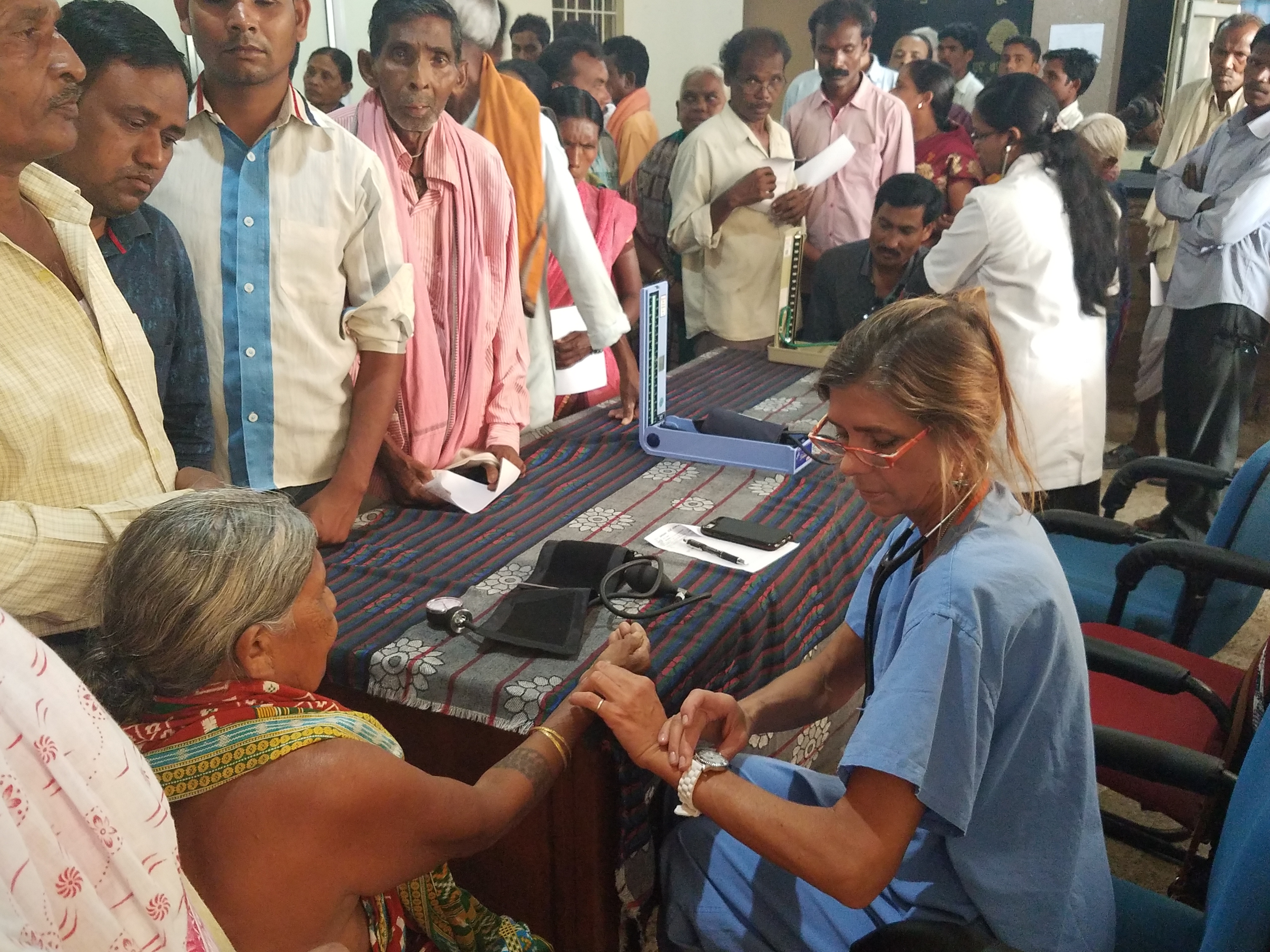 A healthcare worker examines an elderly woman’s hand at a medical camp while a group of people waits in line for checkups inside a busy clinic setting.