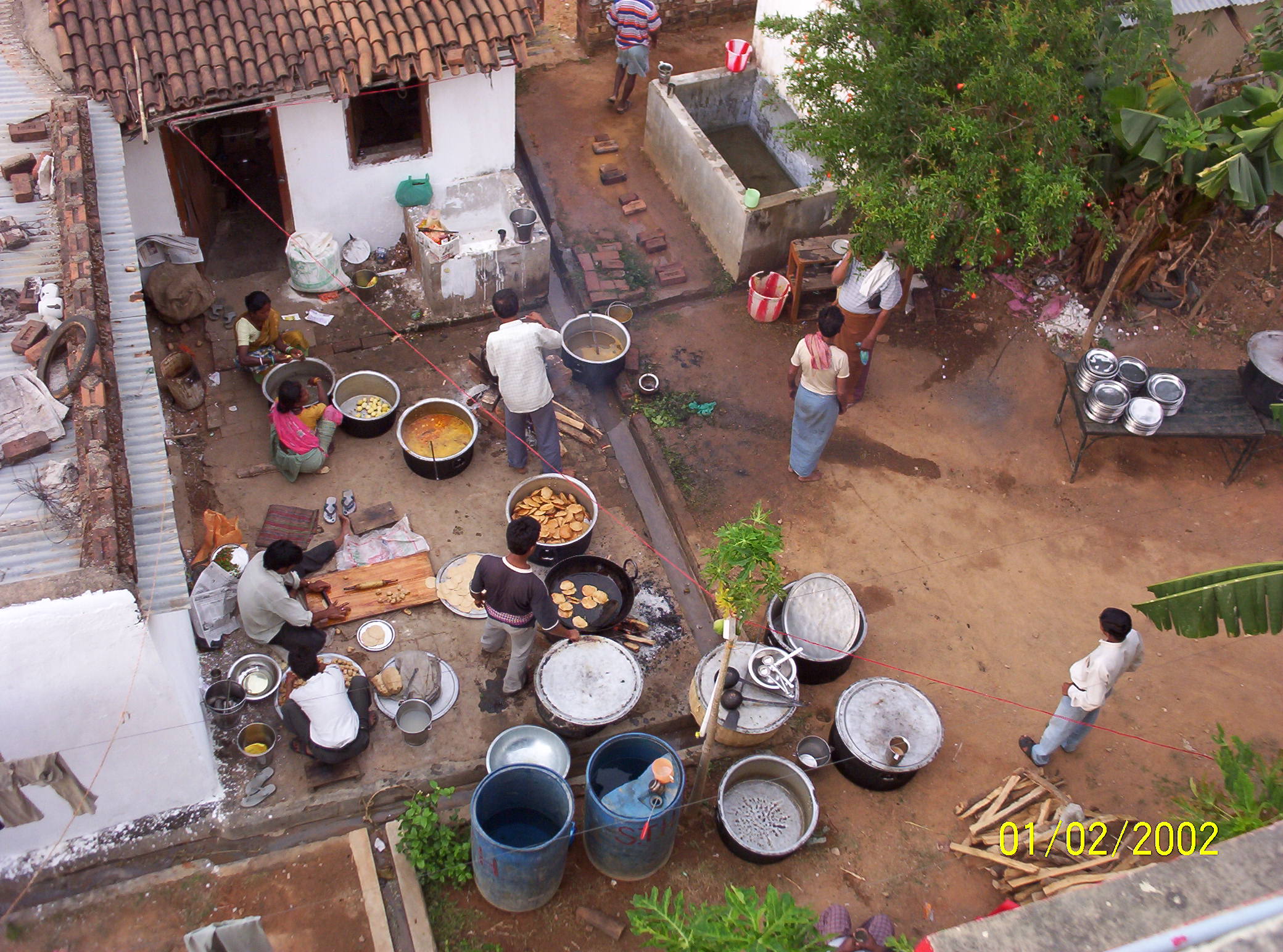 Several people prepare food in large pots and pans in an open courtyard, surrounded by metal containers and a small tiled-roof building.