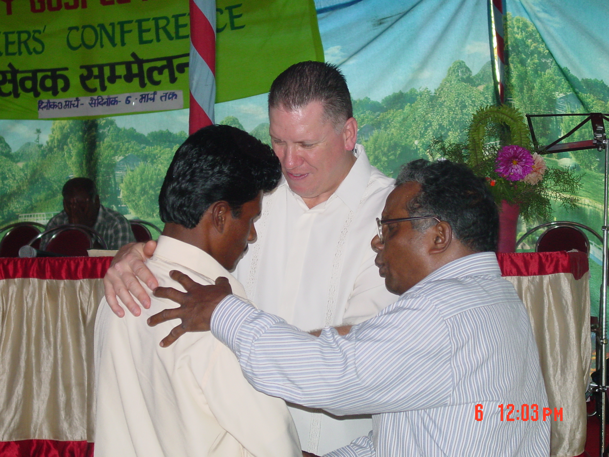 Two men pray with another man on stage during a pastors’ conference in India, with a green banner and floral decorations in the background.