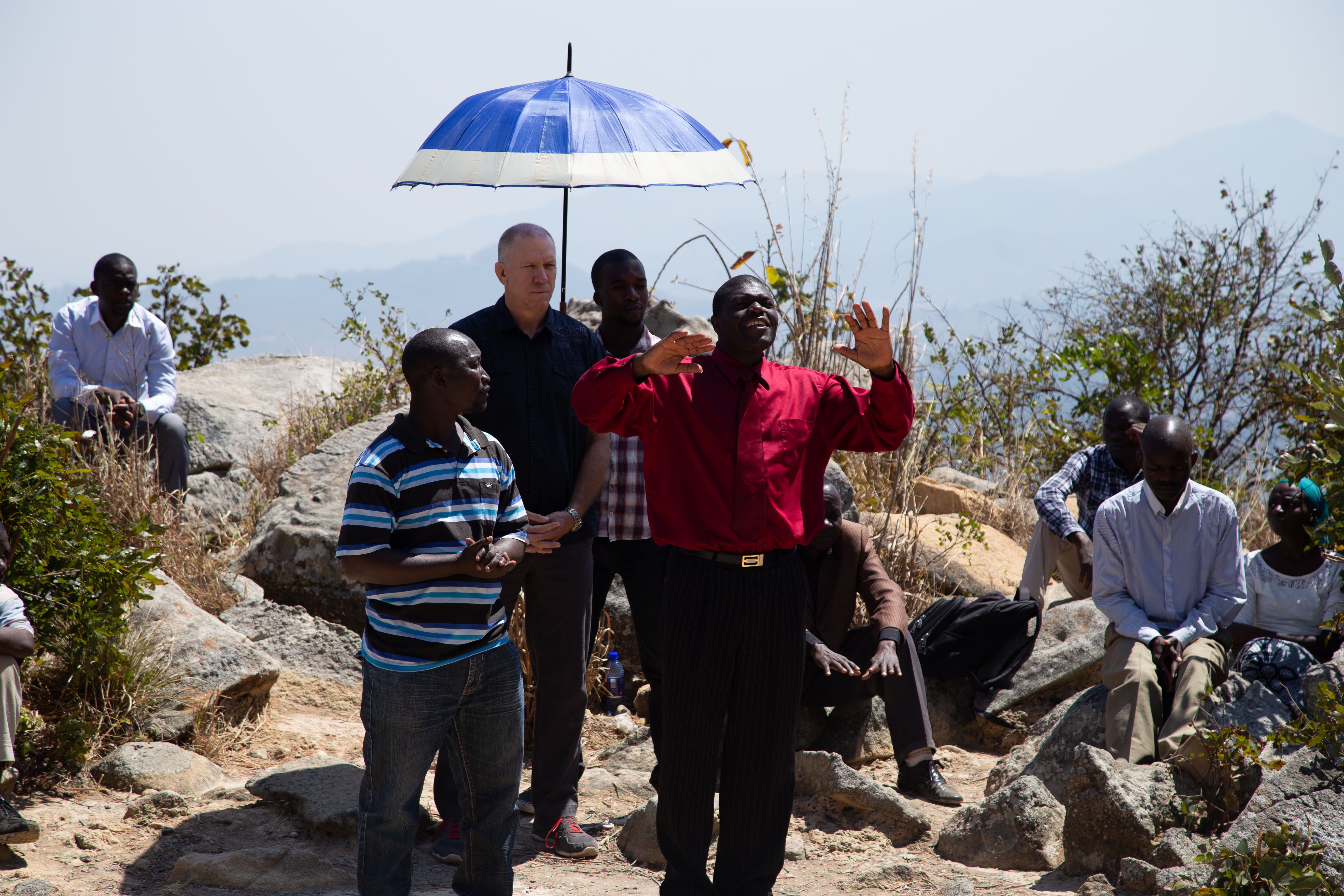A group of men and women meet on a rocky hillside for a prayer or teaching session, with one man in a red shirt speaking passionately under a blue and white umbrella.