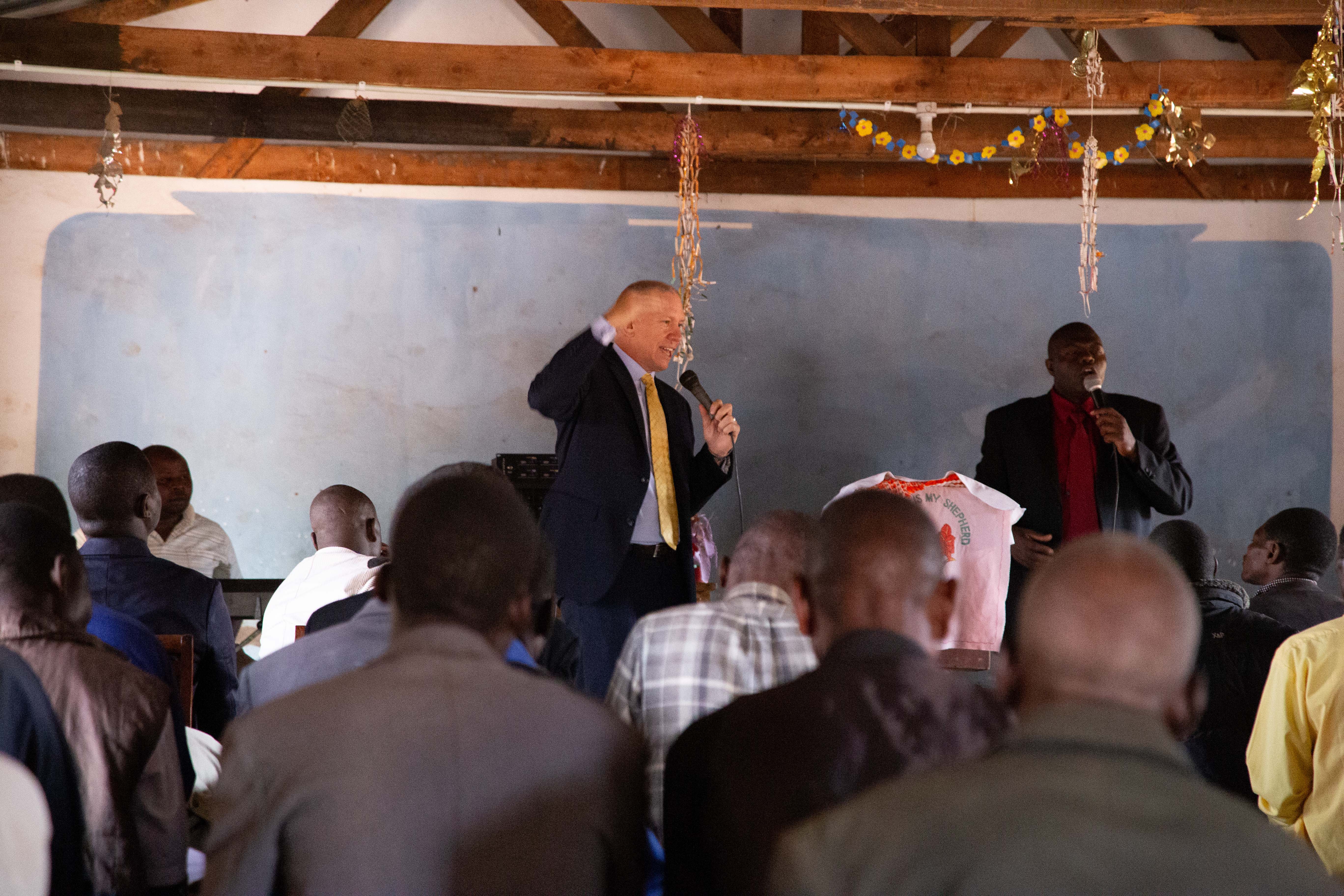 Pastor David prays with church members during an intimate indoor service, with people standing in prayer and colorful clothing visible in the scene.