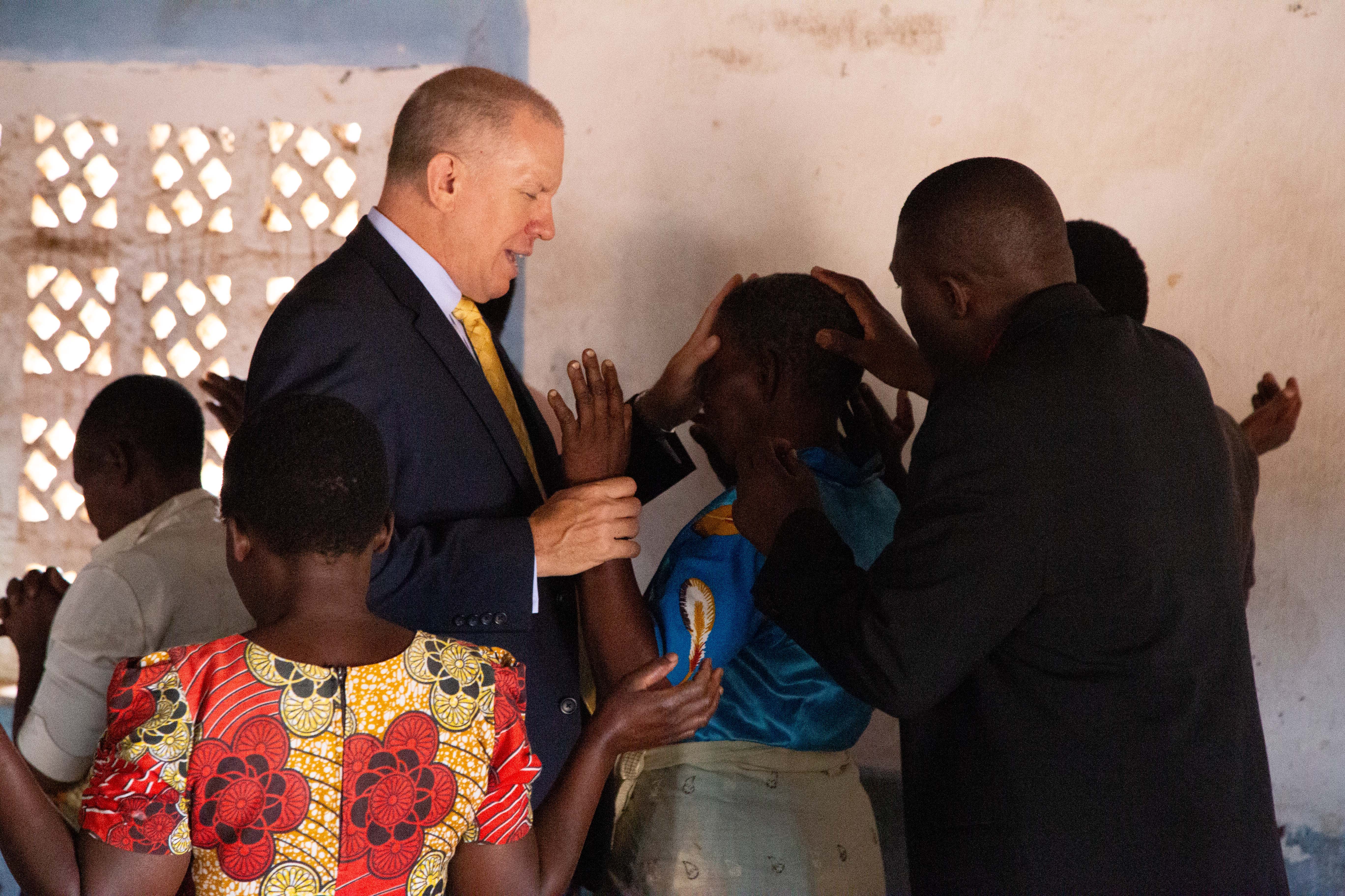 Pastor David prays with members of a congregation inside a simple church building, as several people lay hands and join in prayer.