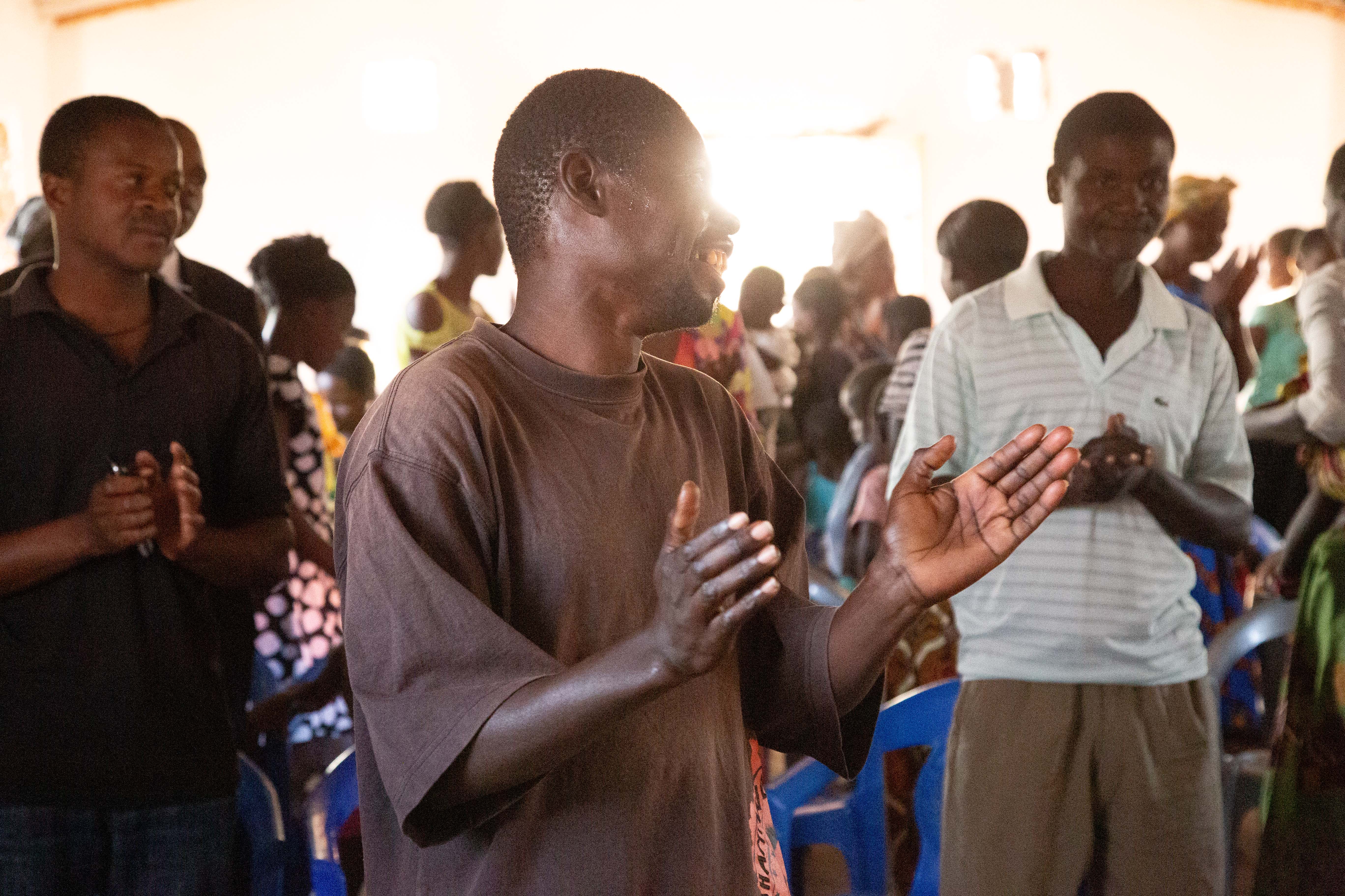 A man claps and sings joyfully among a congregation of people gathered inside a building for worship, with sunlight streaming through the windows.