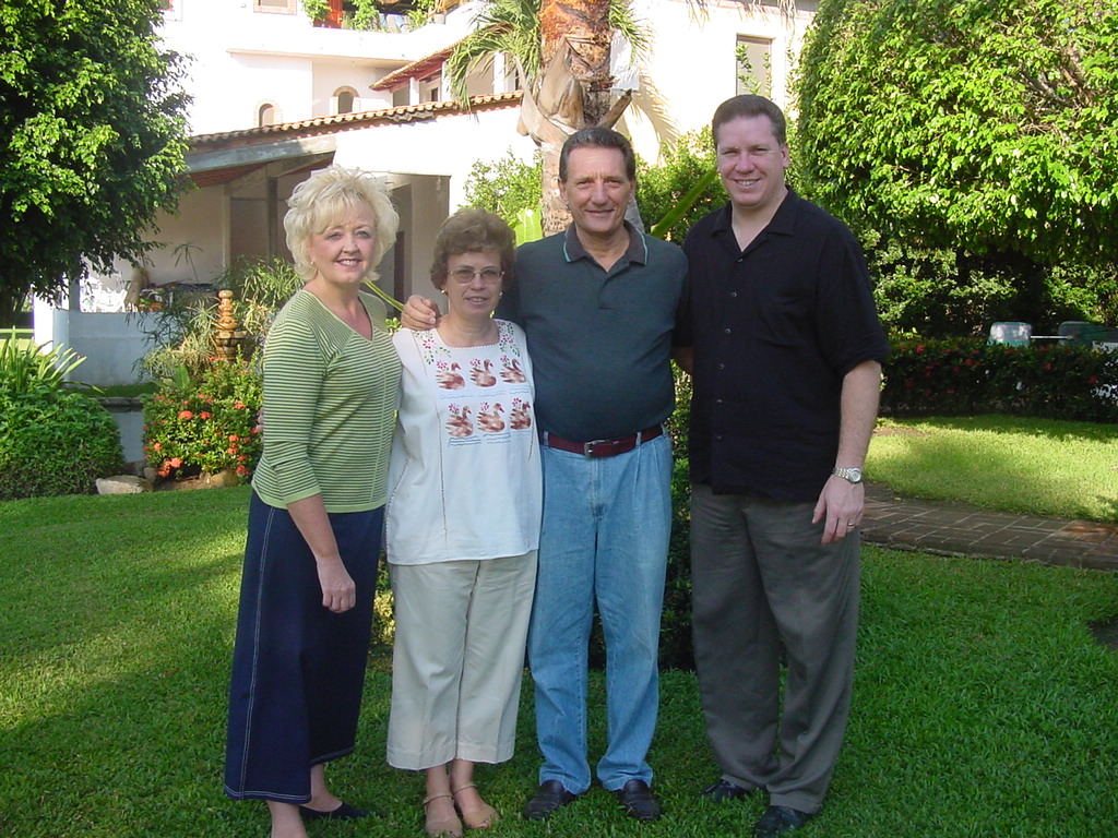 Pastors David, Tracy and friends stand together outdoors in a garden courtyard, smiling for a group photo in front of a building surrounded by trees and flowers.